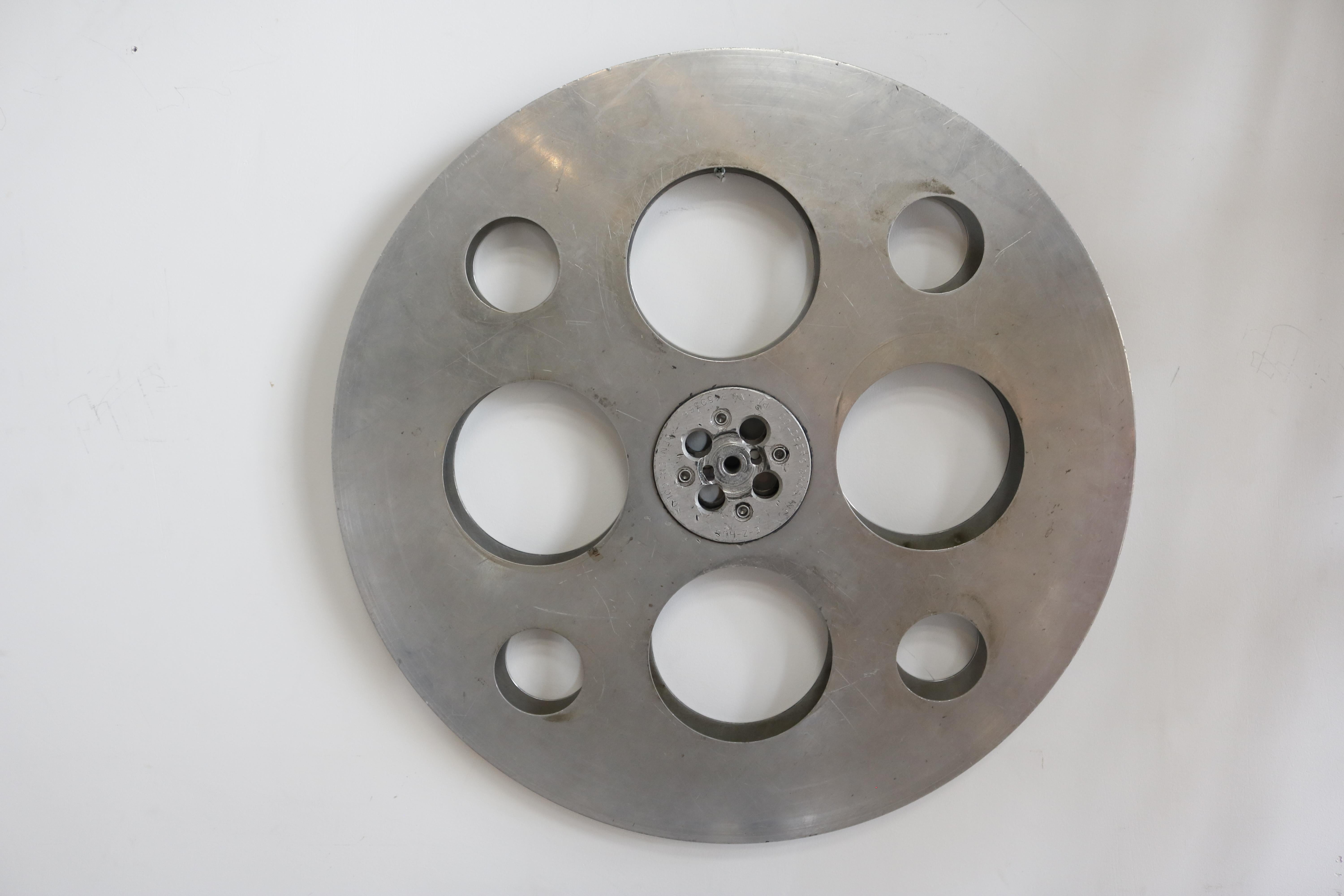 A lustrous, vintage film reel turned wall sculpture made of steel. There are panels held together by a center gear, allowing the them spin. Branded 