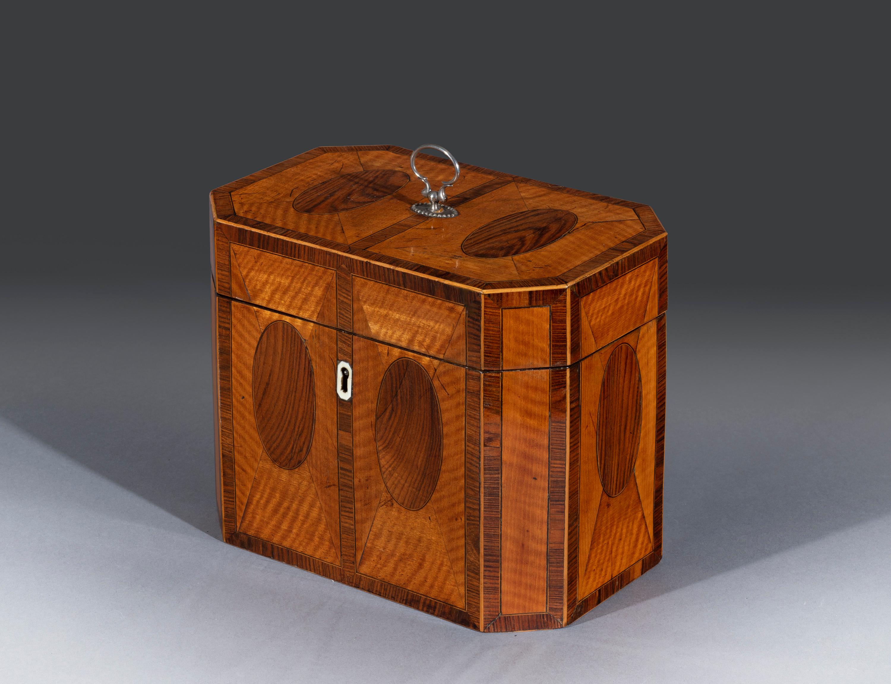The West Indian satinwood caddy is fitted with the original silver handle and is inlaid with padouk and crossbanded and inset with oval panels throughout. The hinged top opens to reveal two glass tea canisters fitted with the original side caps. The