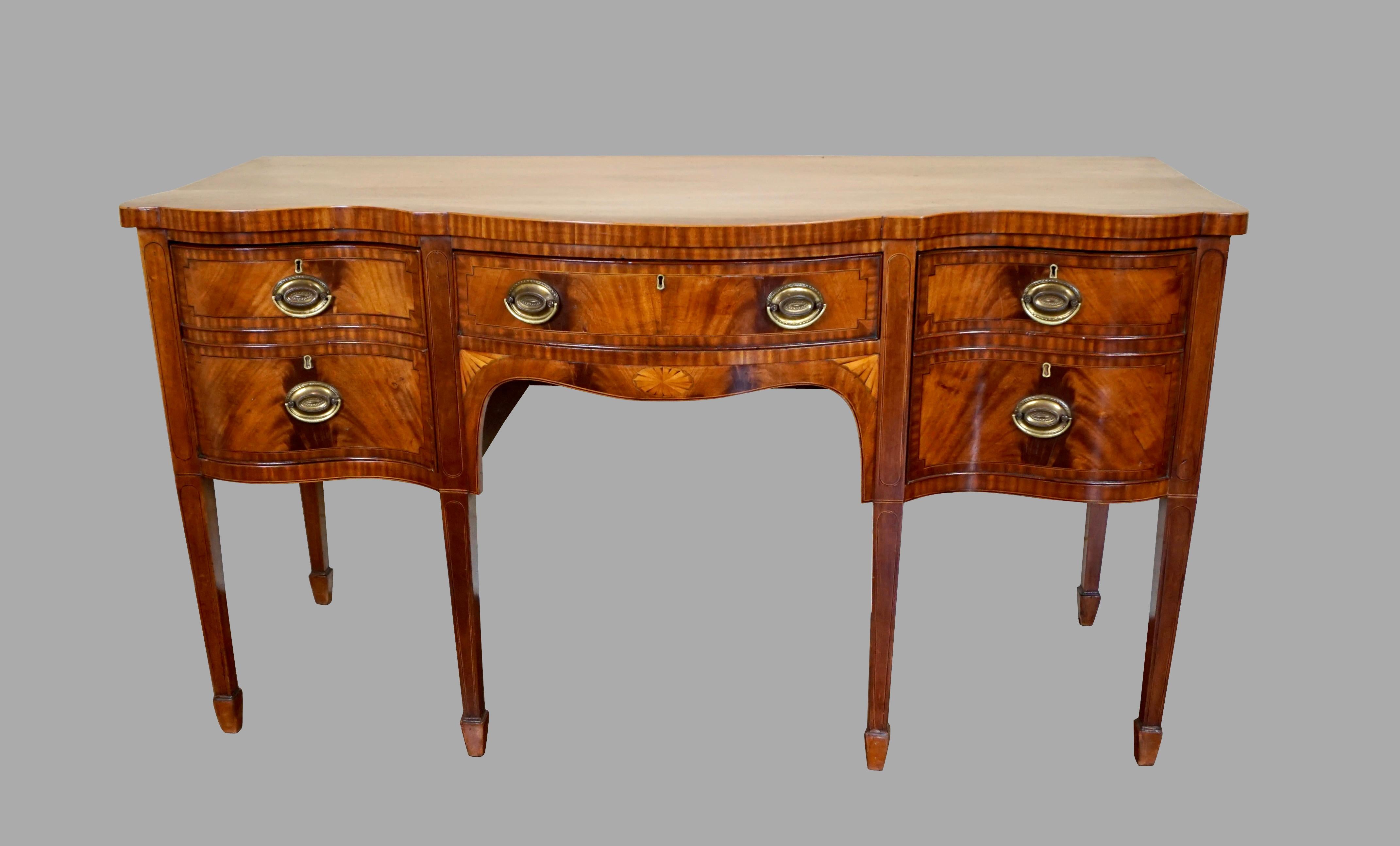 A fine English mahogany satinwood inlaid serpentine front sideboard with crossbanded string inlaid drawers. The center drawer rests above an apron with a satinwood inlaid oval paterae framed with fan inlaid corners. The piece is supported on line