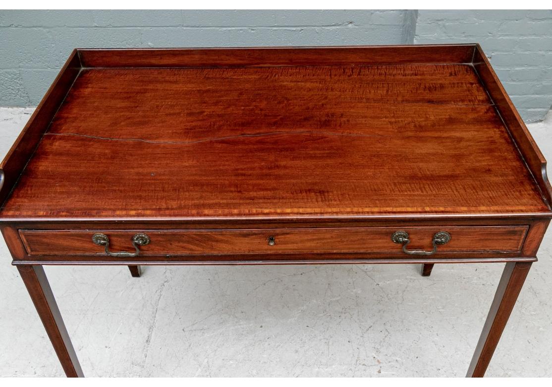 The top with a shaped 3/4 gallery and fine thin banding on the front. The long frieze drawer with the same banding and fine patinated brass bales, flanked by inlaid ovals. Raised on square tapering legs with spade feet. 
Measures: 