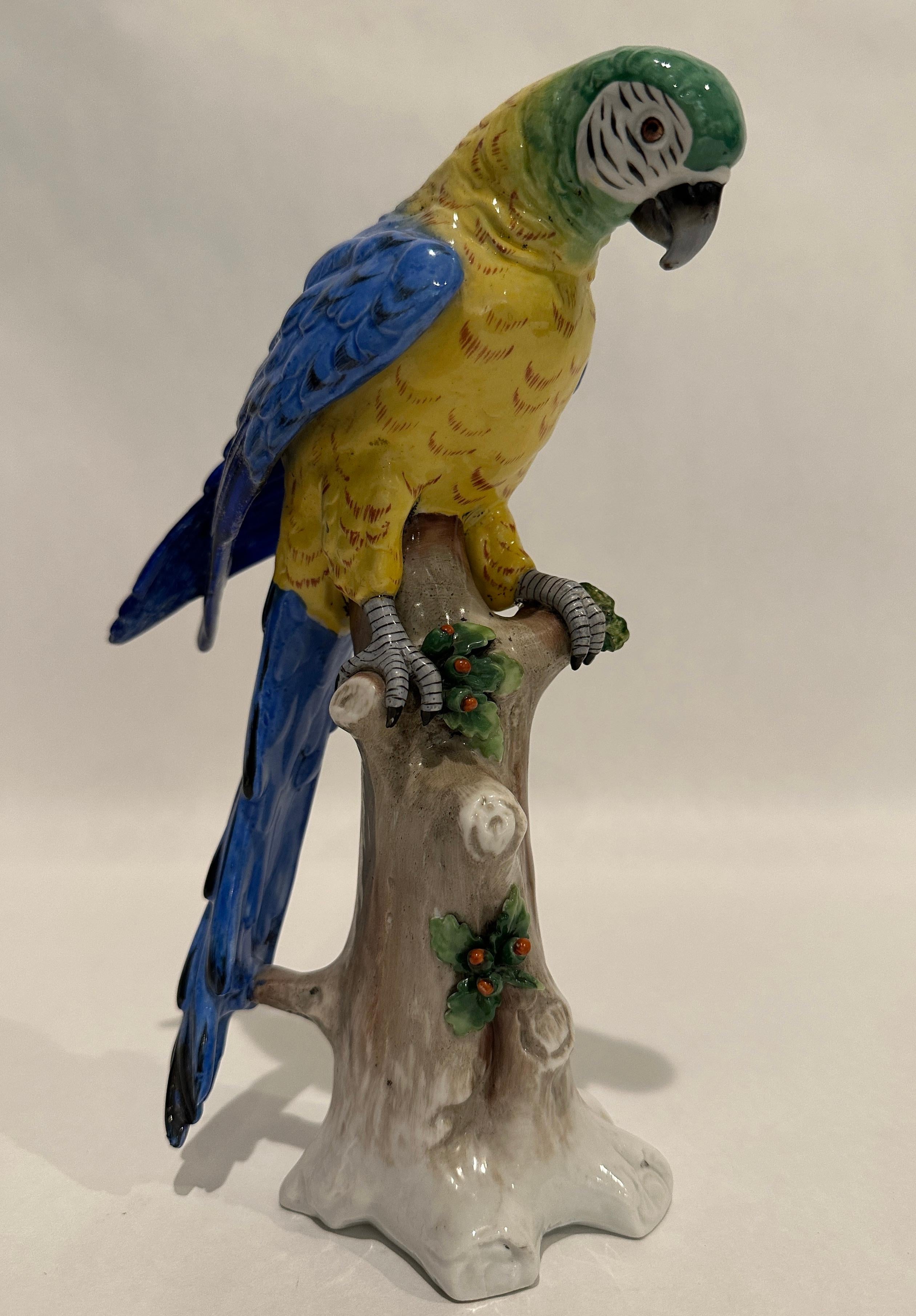 Perroquet en porcelaine coloré et finement peint en vert, jaune et bleu, perché sur une branche réaliste avec des feuilles et des baies. En bas, le 