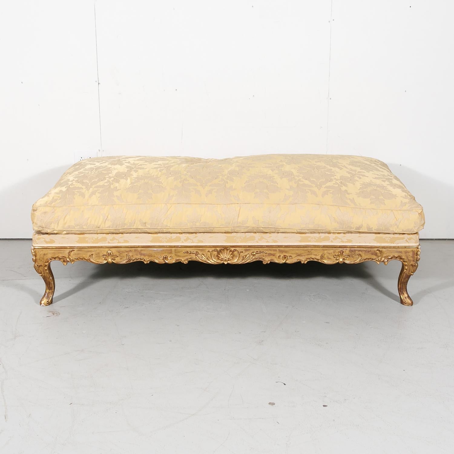A fine 19th century Louis XV style giltwood bench having a loose down cushion upholstered in a beautiful silk damask appliqué atop an elaborately carved upholstered frame with floral and shell motifs on all four sides, 1870s. Raised on short