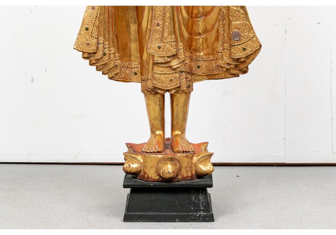 Finely made, the figure stands serenely, dressed in an elaborate pleated robe and headband inlaid with tiny colored mirrors. Finely gilt with patches of red clay visible. The figure stands on a six pointed gilt and red painted base, mounted on a