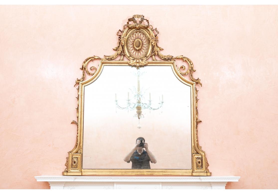 A fine and impressive antique gilt mantle mirror with a large crown comprised of an oval pierced floral motif encompassed by ribbons and acanthus leaves. The scrolling acanthus leaves continue along the shoulders of the mirror encountering carved