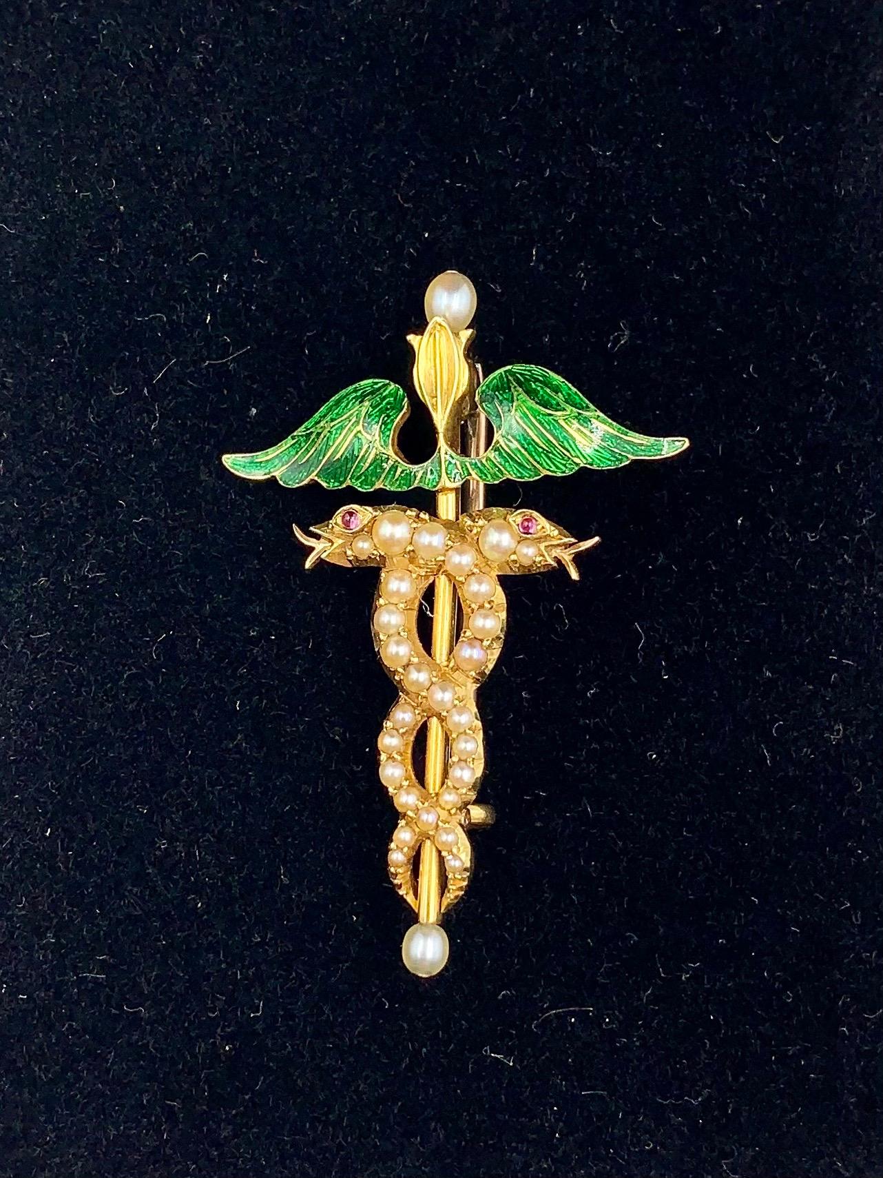  A fine antique Caduceus brooch in 18K yellow gold with a pair of seed pearl entwined serpents with cabochon ruby eyes surmounted by a pair of green guilloche enamel wings centered by a gold staff terminated by two pearls. Beautifully executed