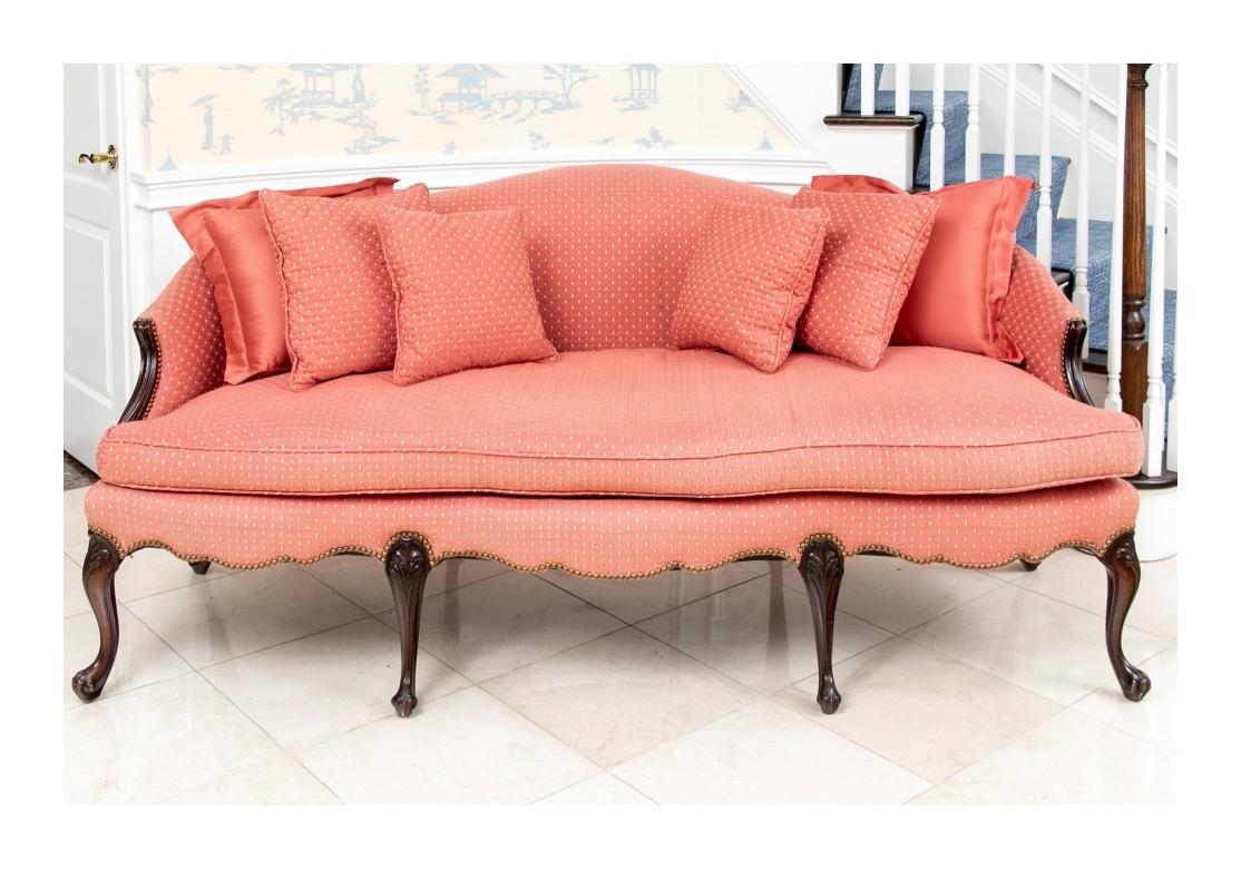 A camel back long sofa, with serpentine seat rail raised on dark stained four cabriole front legs and four curved swept back legs. Tub form sides with sloping wood arms. Upholstered in dusty peach-pink with yellow dotted fabric with nailhead trim,