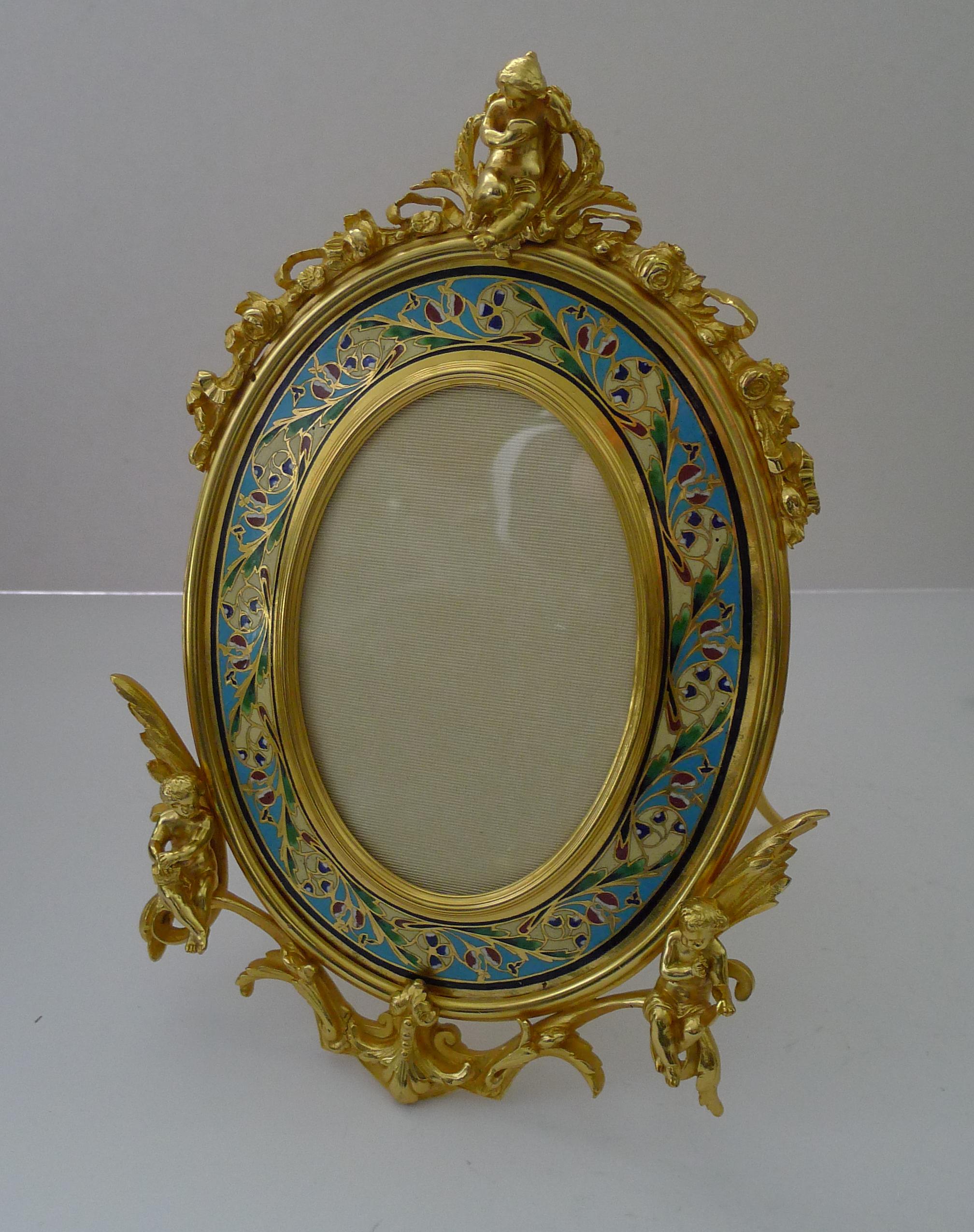 A stunning antique French Bronze Dore photograph frame standing on an easel back stand and also has a ring to the top for wall hanging if desired. The frame is decorated with champleve enamel and mounted with three stunning Cherub or Putti