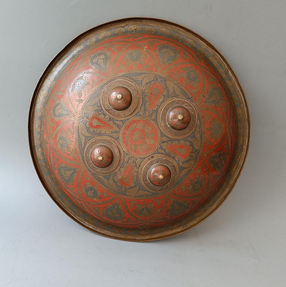  
 A antique Indian Dhal shield made from hammered brass, circular in  form with an curved outer rim and four dome peaks connected to 4 metal rings in the interior  connect to  cloth hand straps.  The exterior decorated with hand chiselled and