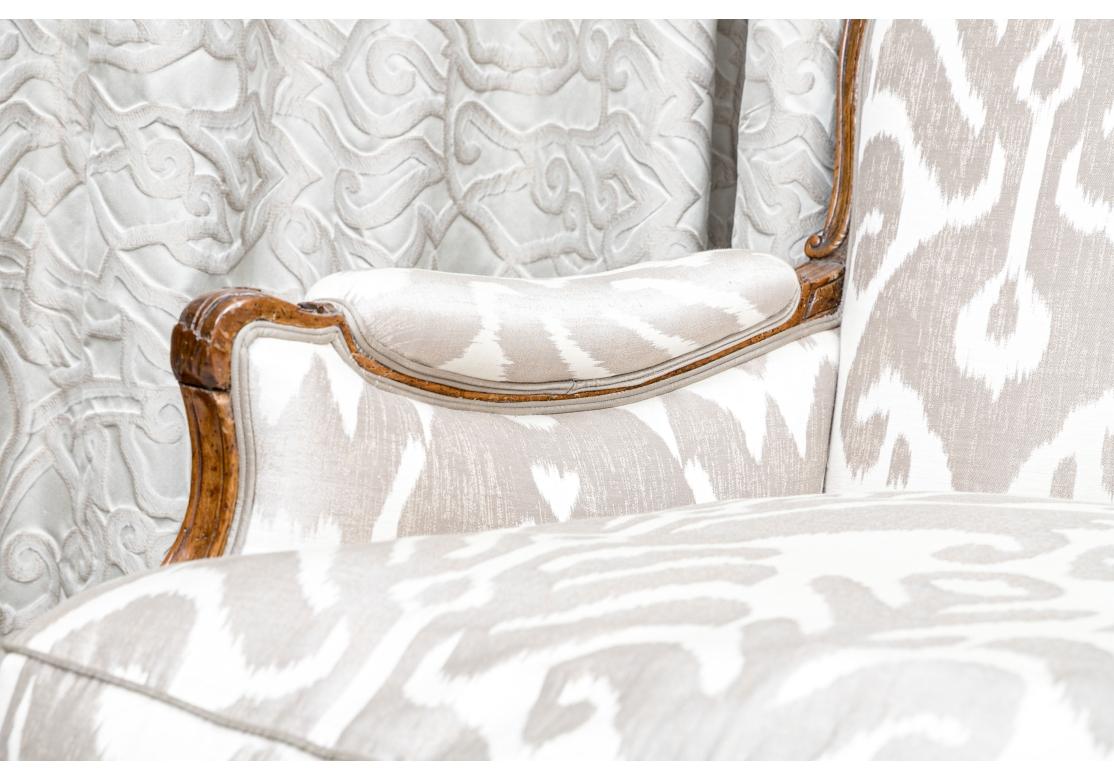 A fine Louis XV style upholstered bergere with deep seating and carved fruitwood frame, floral cresting rail and resting upon carved cabriole legs. The chair is upholstered in a patterned two Silk fabric with sheen and solid piping. The side panels
