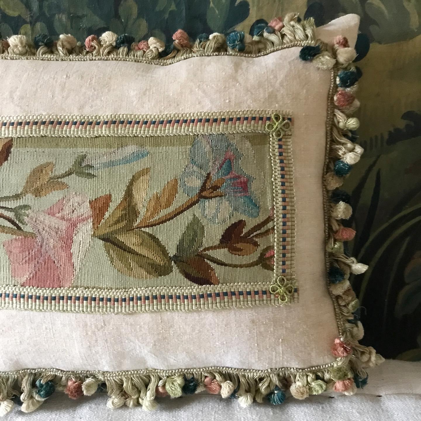 A very beautiful cushion made from a panel of 19th century French Aubusson placed onto a pale sandy coloured antique hemp linen then trimmed with a complimentary colour matched braid with hand made rosettes on each corner then a matching thick fan