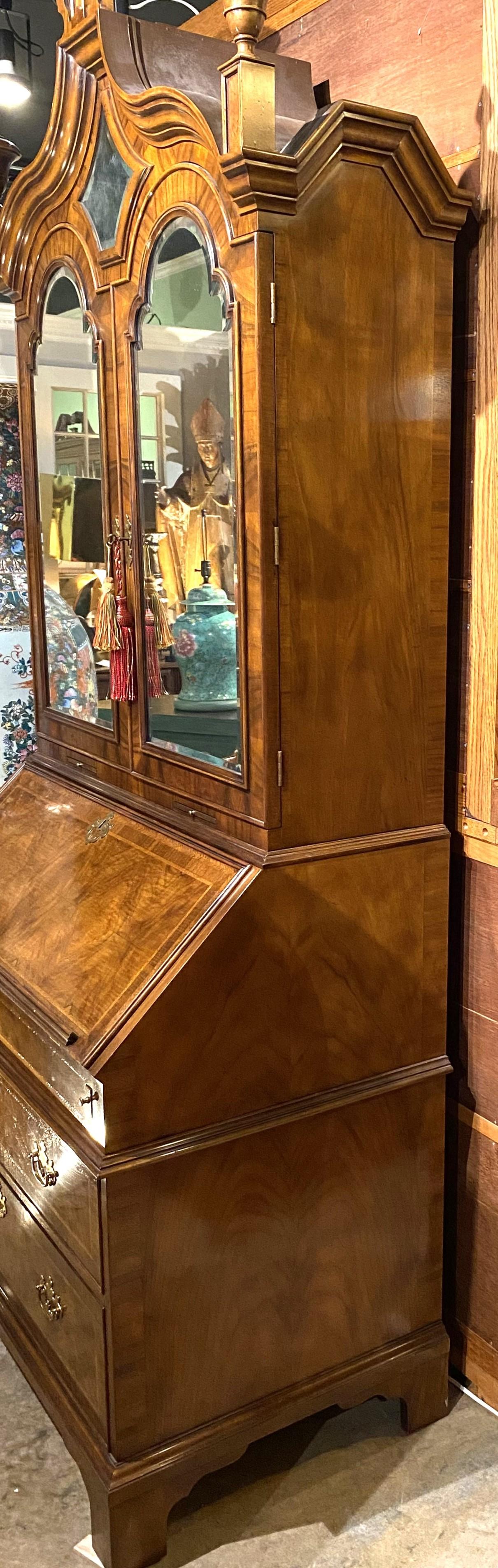An exceptional Georgian style two piece inlaid secretary in burled walnut by Baker Furniture, its upper case with a shaped crest with center mirror adorned with three urn finials surmounting two arched beveled mirrored doors, opening to a multitude