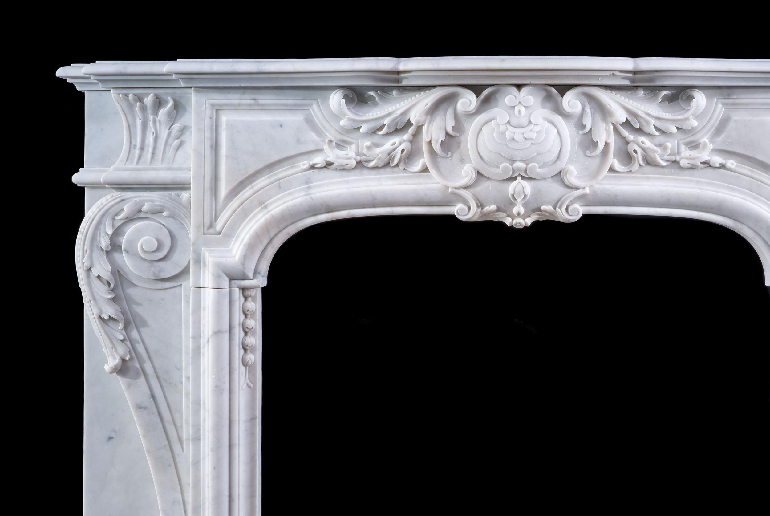 A small but grand Belgian fireplace in softly mottled Carrara marble. The moulded shelf sits above a panelled frieze which is centred by a very ornate foliate cartouche, and is flanked by broad acanthus console jambs over plain staggered