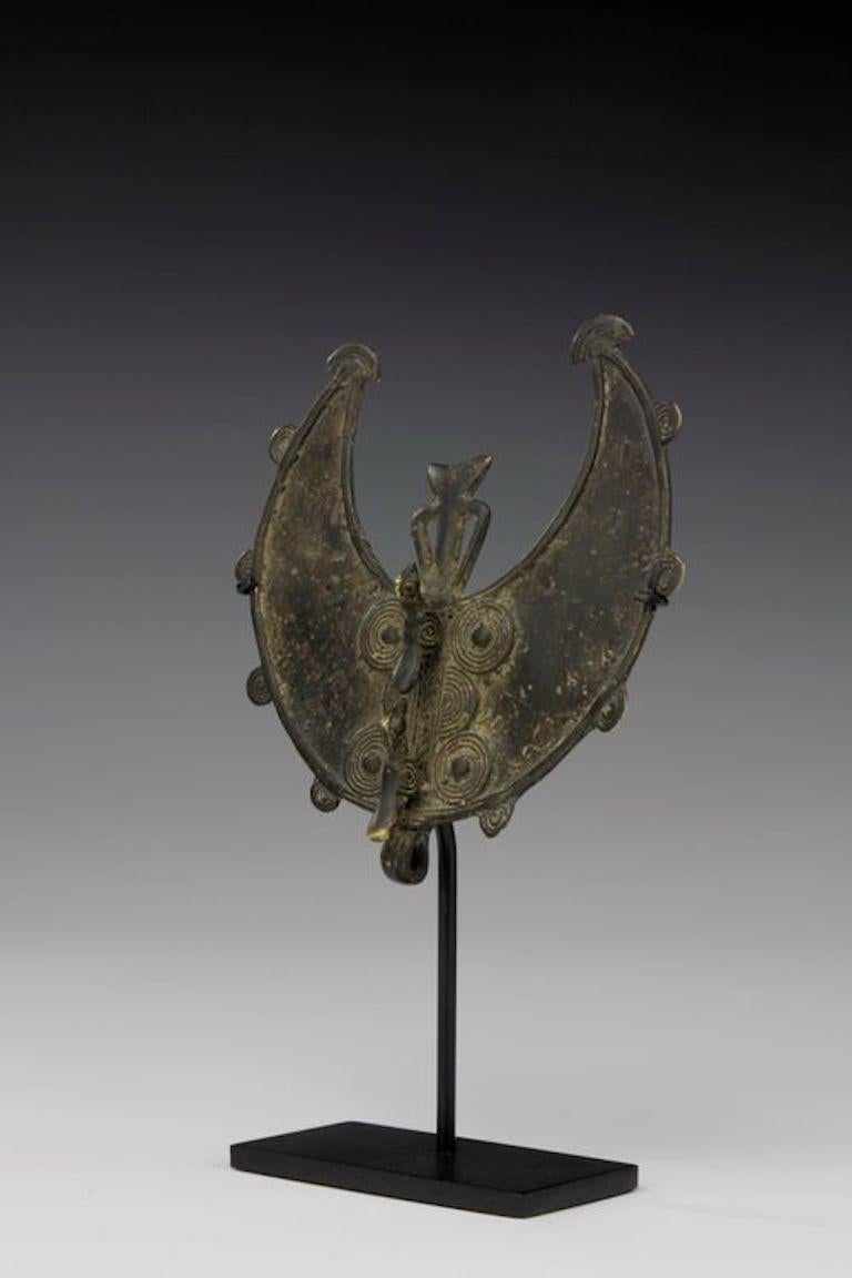 Este colgante de bronce en forma de media luna, presenta una figura abstracta central. Debajo de la figura, dos pequeños pájaros estilizados se posan en el centro del colgante. En toda su superficie, el colgante de bronce muestra una preciosa pátina