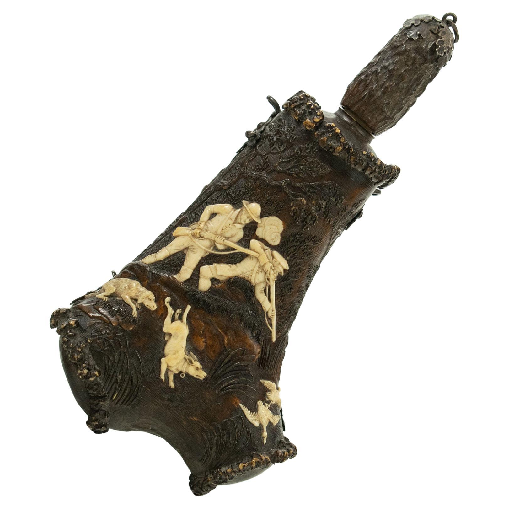 A FINE CARVED GERMAN OR AUSTRIAN 18TH CENTURY STAGS HORN POWDER FLASK - The front face expertly carved with two hunters in a wooded river bank scene with hounds and two flighting ducks, screw cut, bronze oak leaf mounted cap, turned horn nozzle and