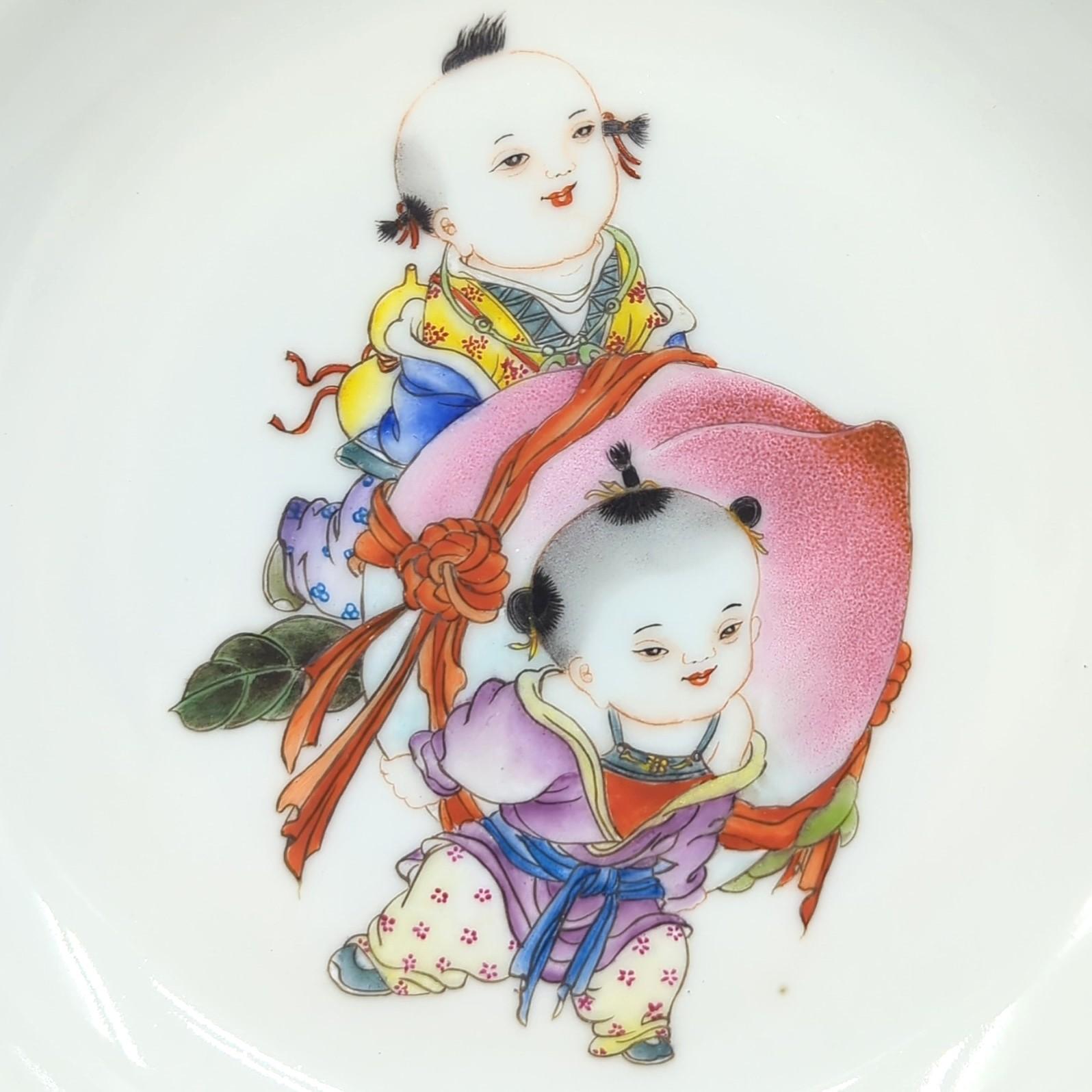 This Chinese porcelain plate, dating from the early 20th century Republic period, is a remarkable representation of both artistic skill and cultural symbolism. The plate features a meticulously executed scene of two boys in traditional Chinese