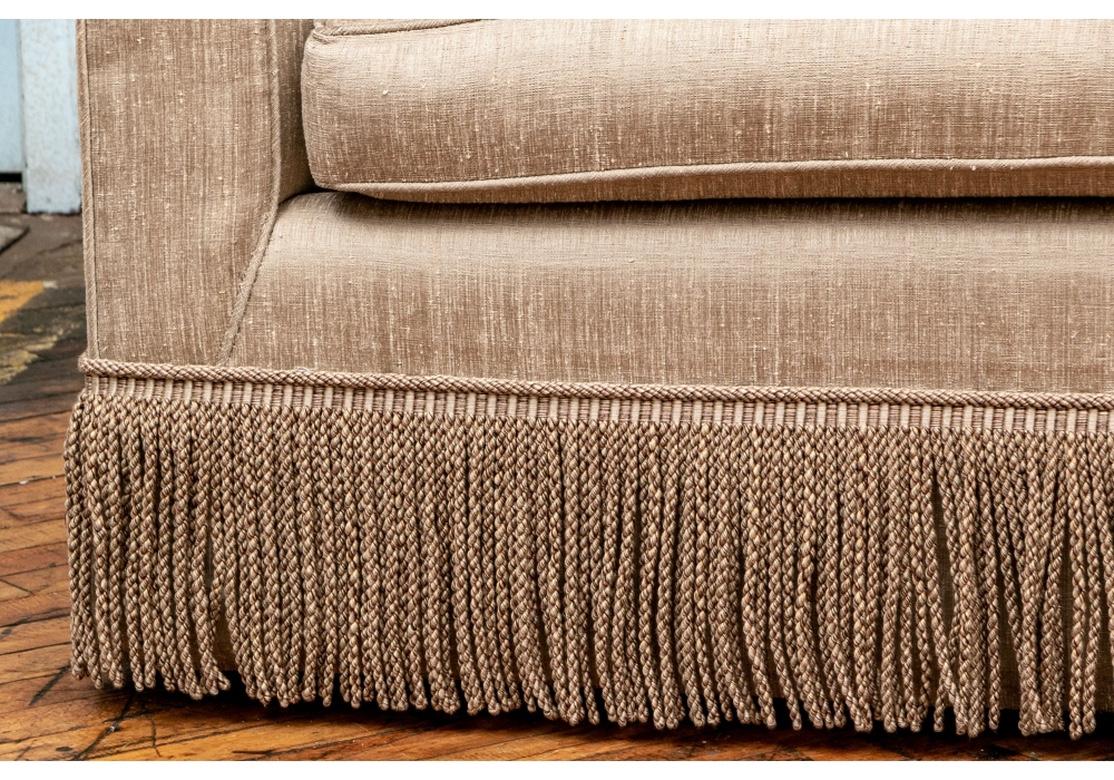 The sofa with square lines, a slightly shaped crest rail and tall sides. With a single seat cushion and fringe around the skirt. Upholstered in a fine nubby silk blend fabric in a great neutral pale Champagne tone and coordinating fringe. From the D