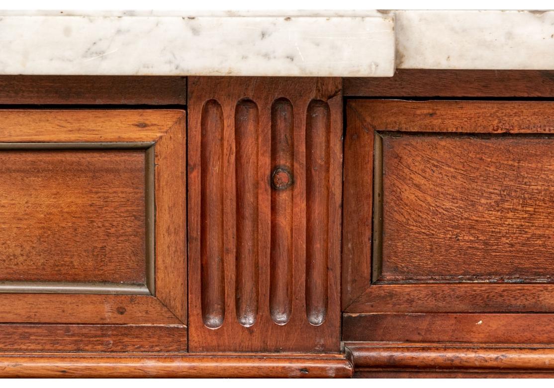 A tall antique French sideboard in mahogany with canted corners and white marble on top.  With three recessed flame frieze drawers with carved moldings, each with a decorative brass escutcheon with garlands and bows, with one key. The case with