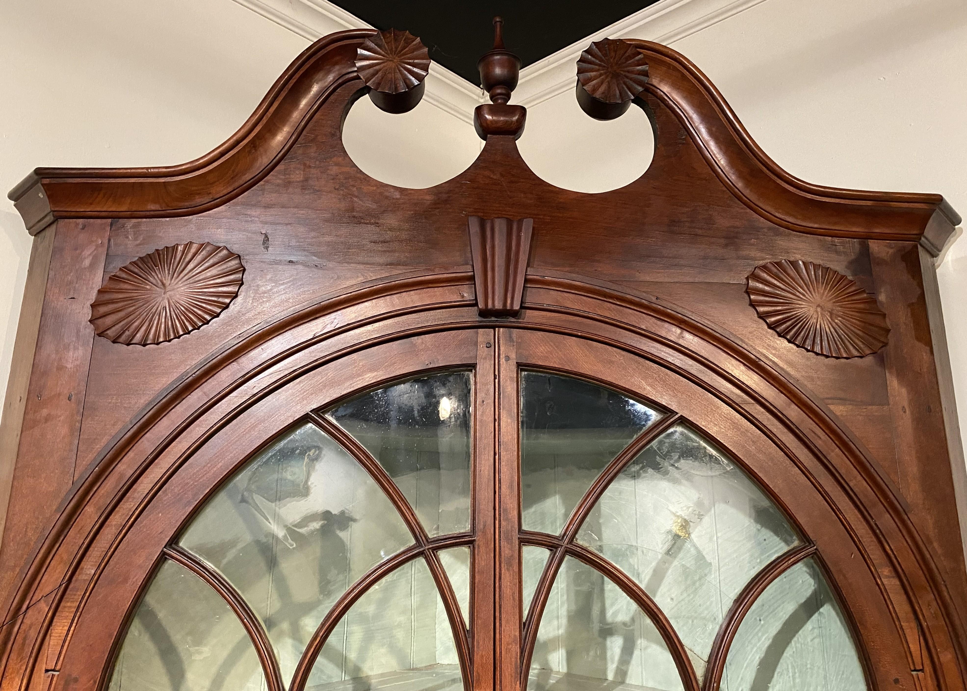 Un'imponente credenza angolare in due pezzi in pioppo e pino, la cui cassa superiore presenta uno stemma a frontone spaccato a collo di cigno con terminale centrale a urna affiancato da due rosette intagliate che sormontano due porte ad arco con