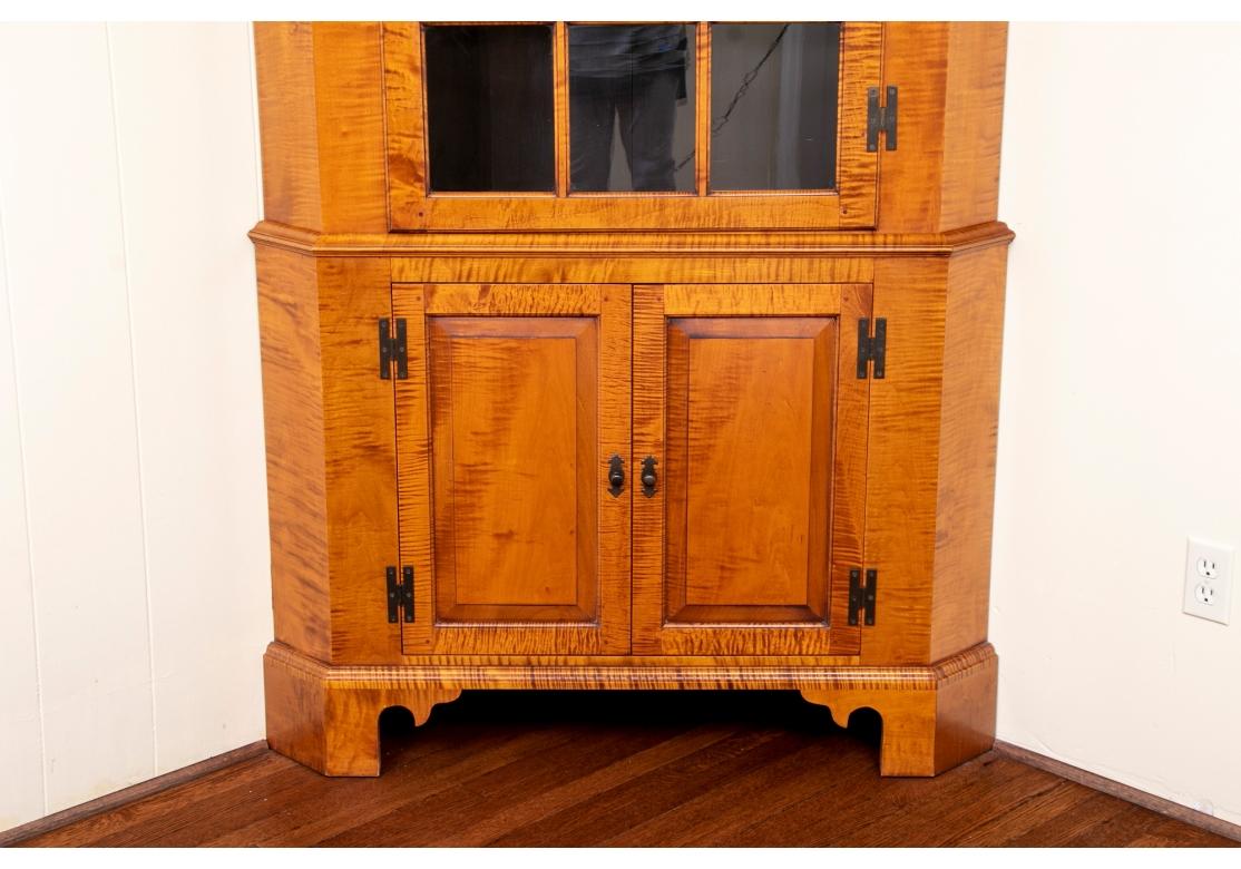 Eldred Wheeler tiger maple corner cabinet featuring individual pane glass, pegged construction, and brass hardware.  Comes in two pieces for ease of transport and placement. Upper case with molded cornice above a single 12 glazed panel door with