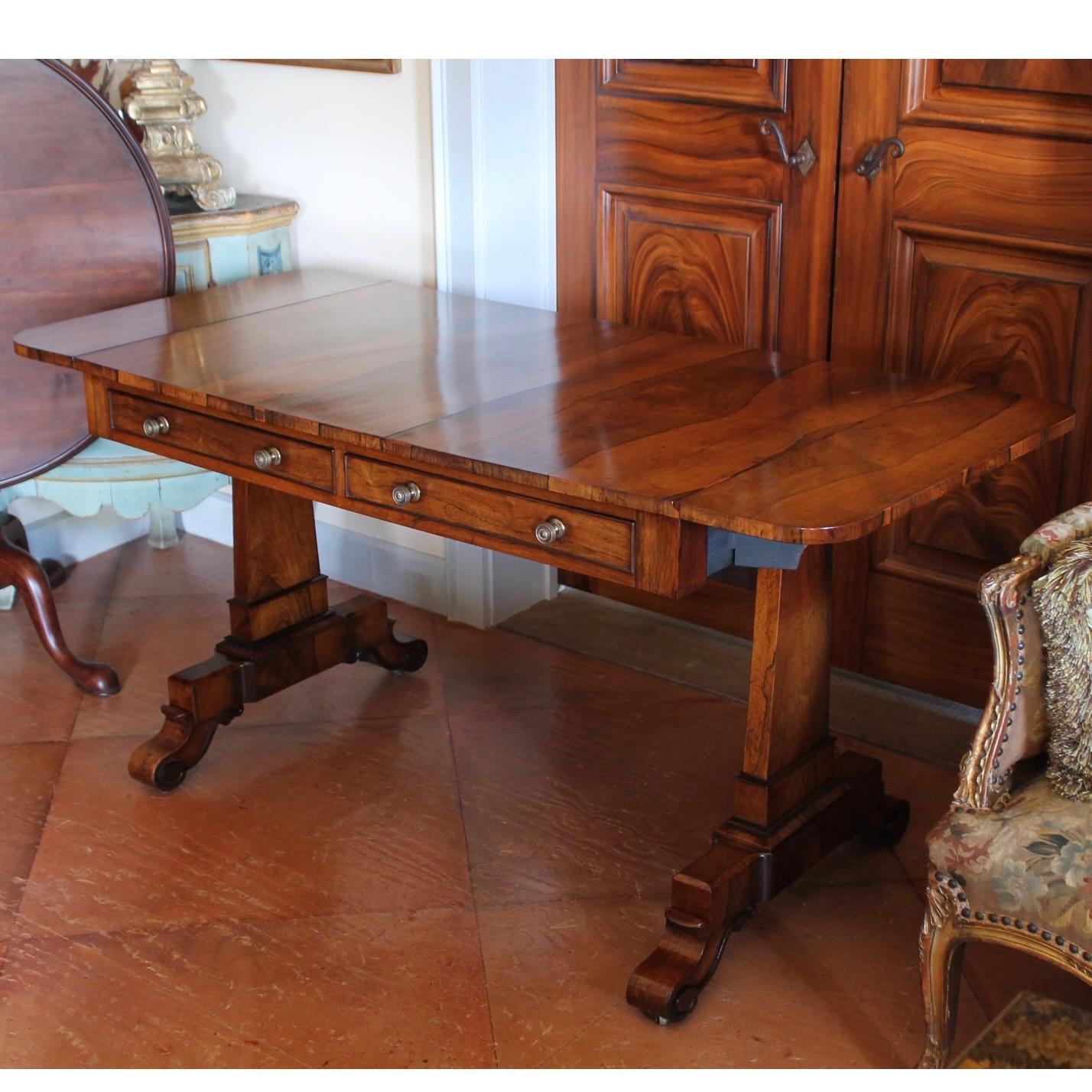 A striking and elegantly contoured sofa table resting on slender obelisk form pedestal legs supported by carved scroll feet on casters. Two functional drawers on one side and with false drawer fronts on the reverse. There are two narrow drop leaves