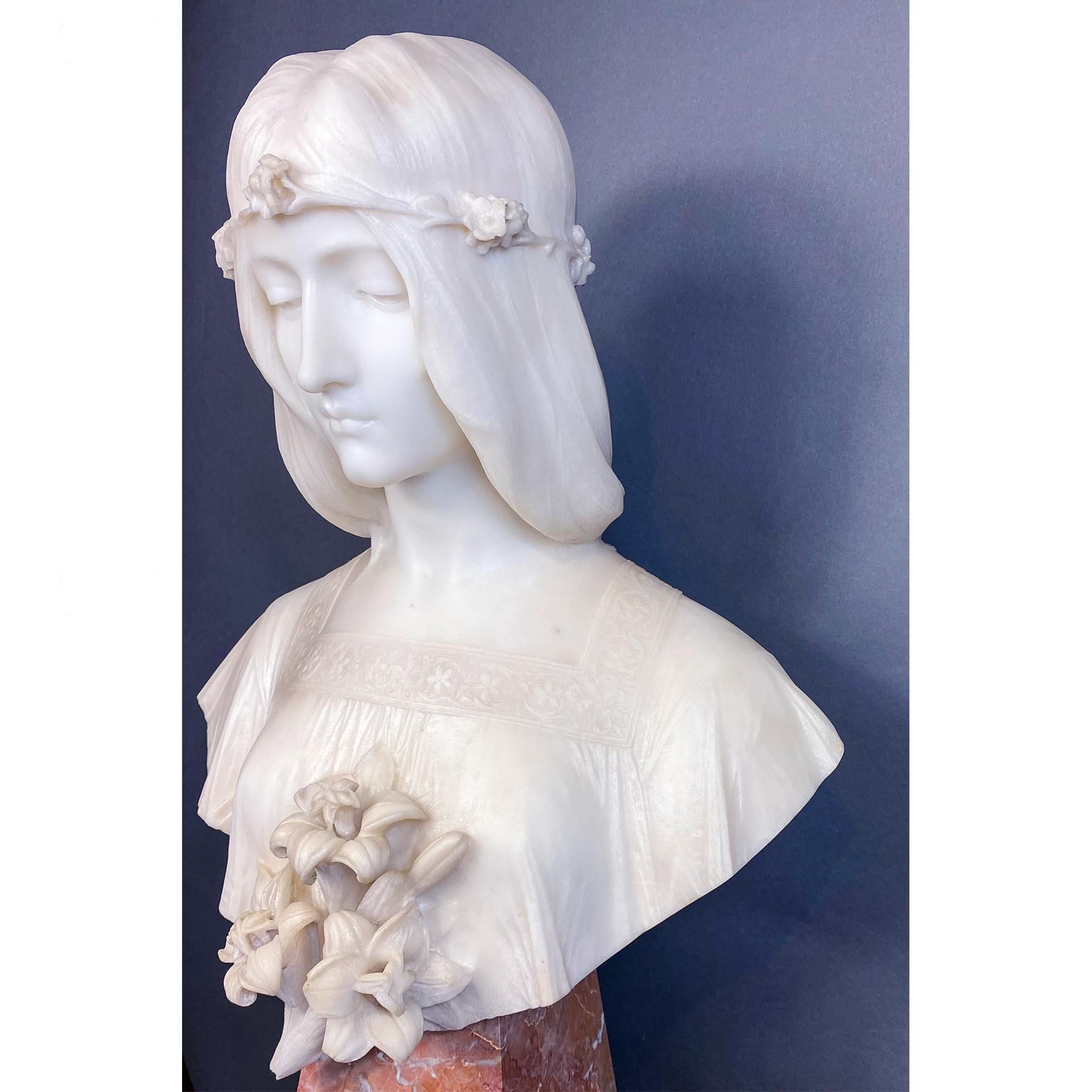 Magnifique buste féminin portant un haut brodé à encolure carrée et une couronne de fleurs. Un bouquet de lys orne le devant du buste qui repose sur une base carrée effilée en marbre contrastant. De délicates draperies sont habilement modelées par