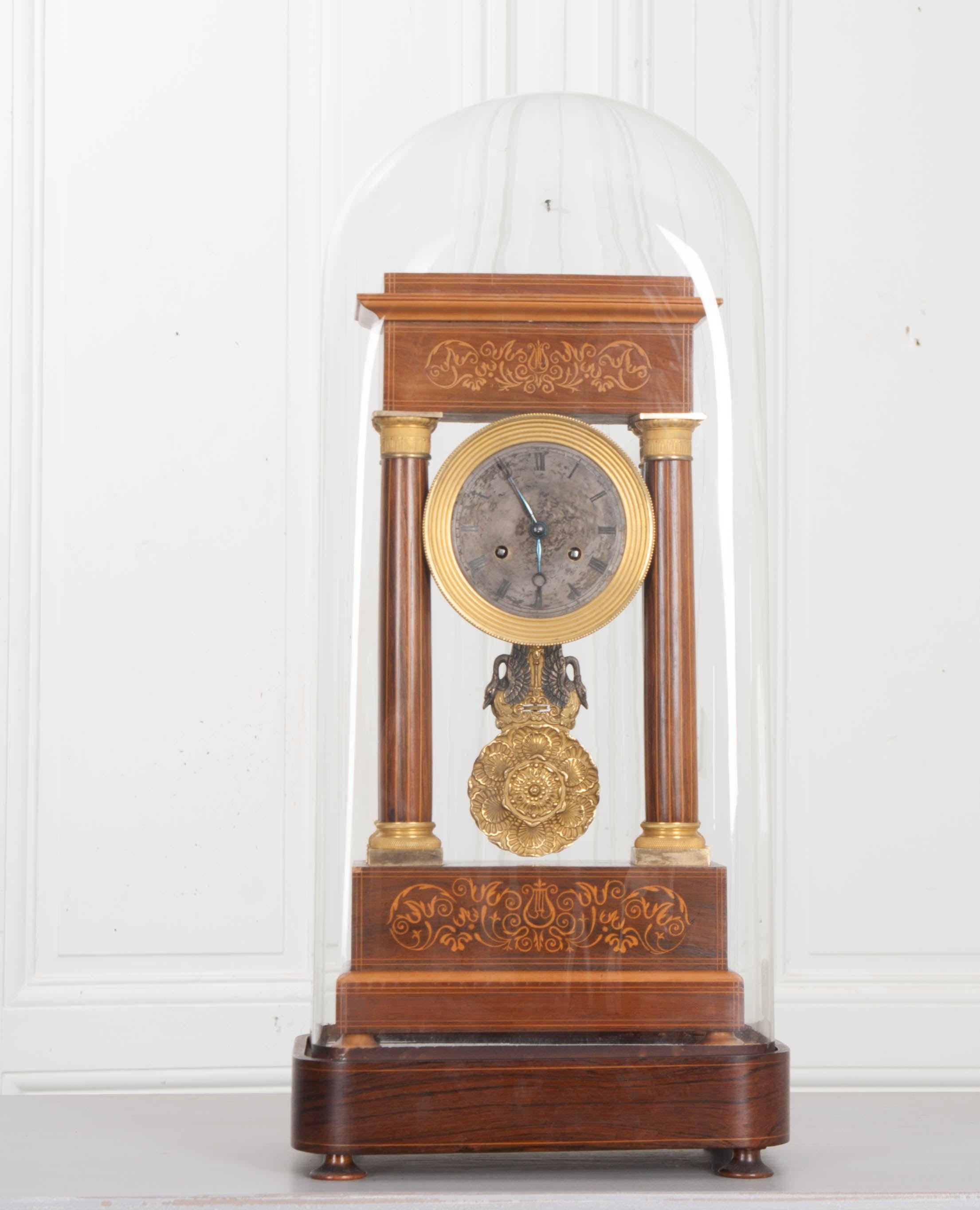 Cette horloge portique française du XIXe siècle en marqueterie de satin et bronze doré, vers les années 1840, présente une frise en marqueterie de fleurs et de folioles sur quatre colonnes en satin marquetées au trait, avec des chapiteaux en bronze