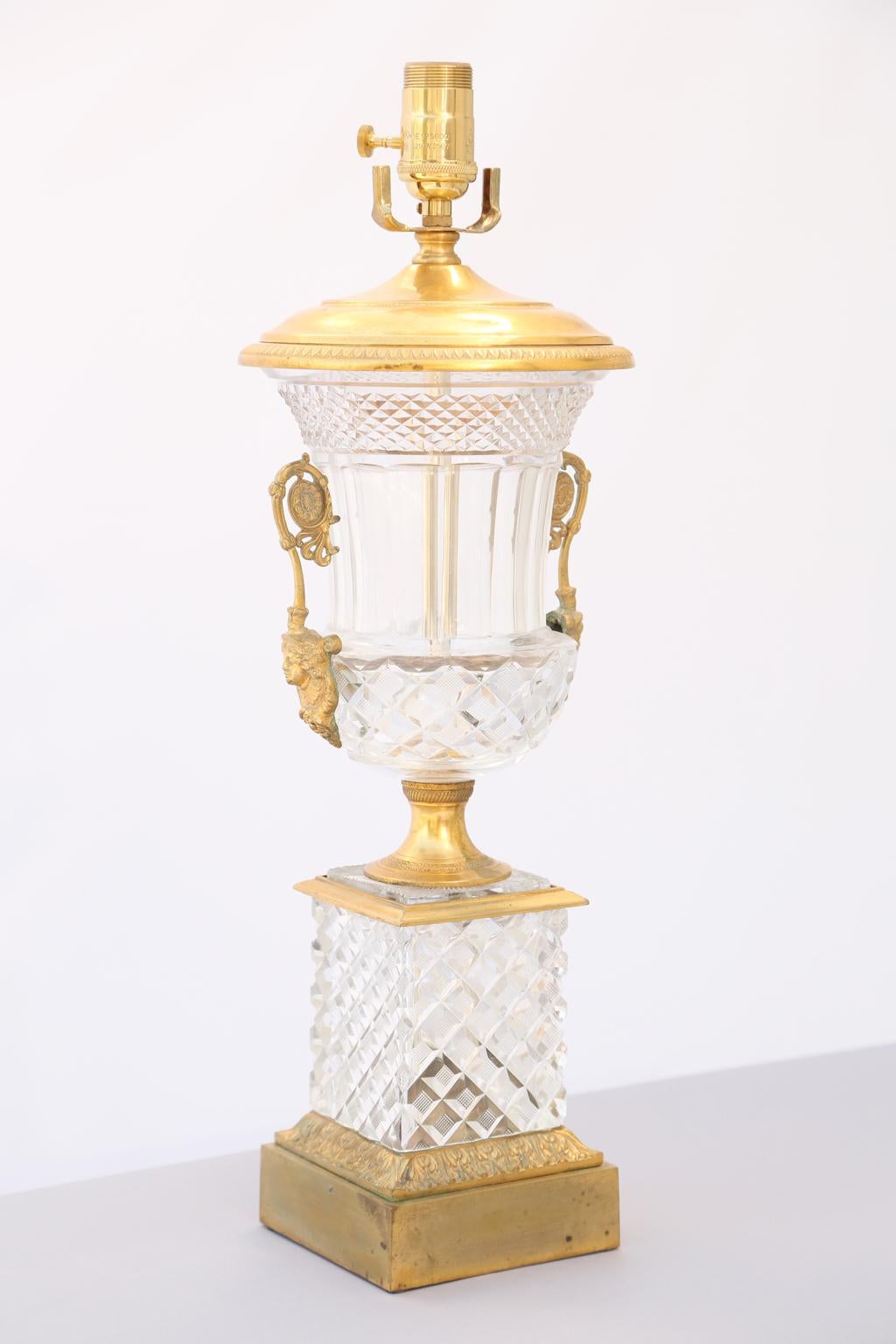 A lamped, campana urn vase, of lead crystal and bronze doré, its upturned diamond-cut lip, over sloped neck, to crosscut-diamond body, flanked by scrolling handles, centred with a rosette, and ending on a classical female mask-mount, raised on gilt