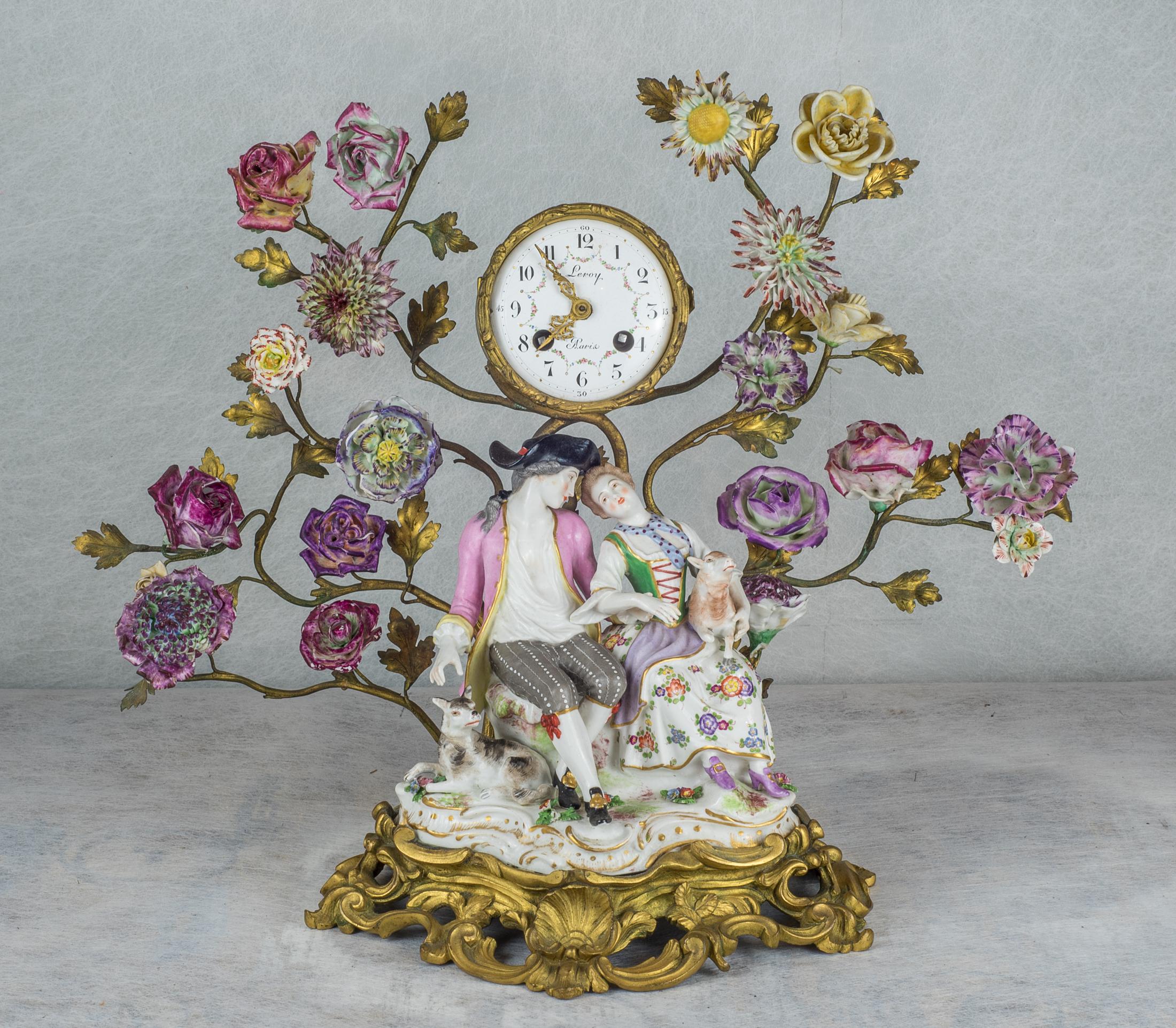 A fine French gilt-bronze and porcelain clockset with figural group and porcelain flowers.

The figural group modeled as 18th century lovers, the woman carrying a lamb, a dog beside the gentleman, another gentleman playing a flute with a dog
