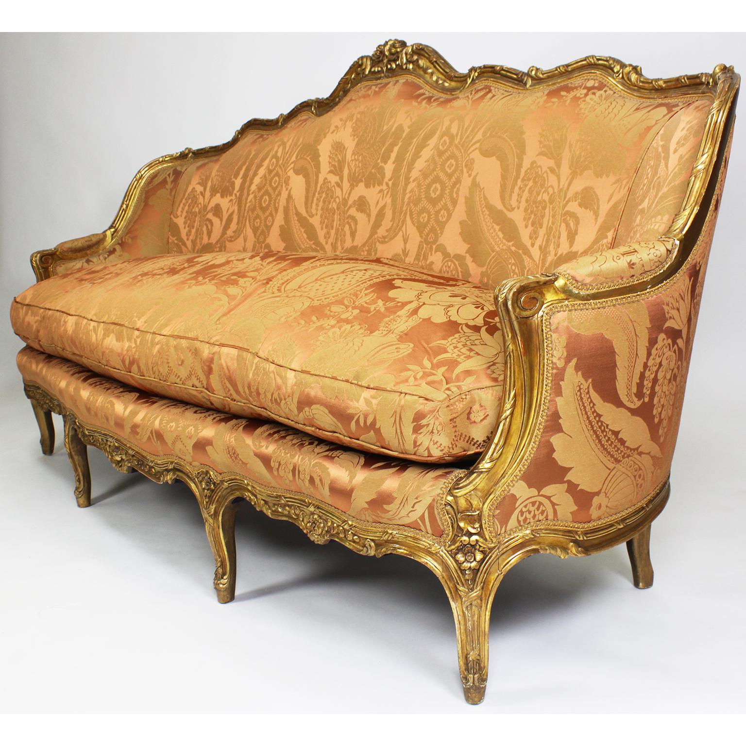 A fine French Louis XV style giltwood carved bergère settee (sofa). The serpentine carved gilded frame crowned with floral design, eight cabriolet legs with a padded backseat, sides and back, with a large cushion pillow seat upholstered in a recent