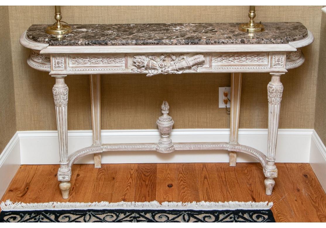 A striking carved wood console with white washed console table with polished and beveled marble top. The Demi Lune form carved apron with a central heraldic device of Torch and Quiver with Flower Garland resting on fluted and tapering legs conjoined