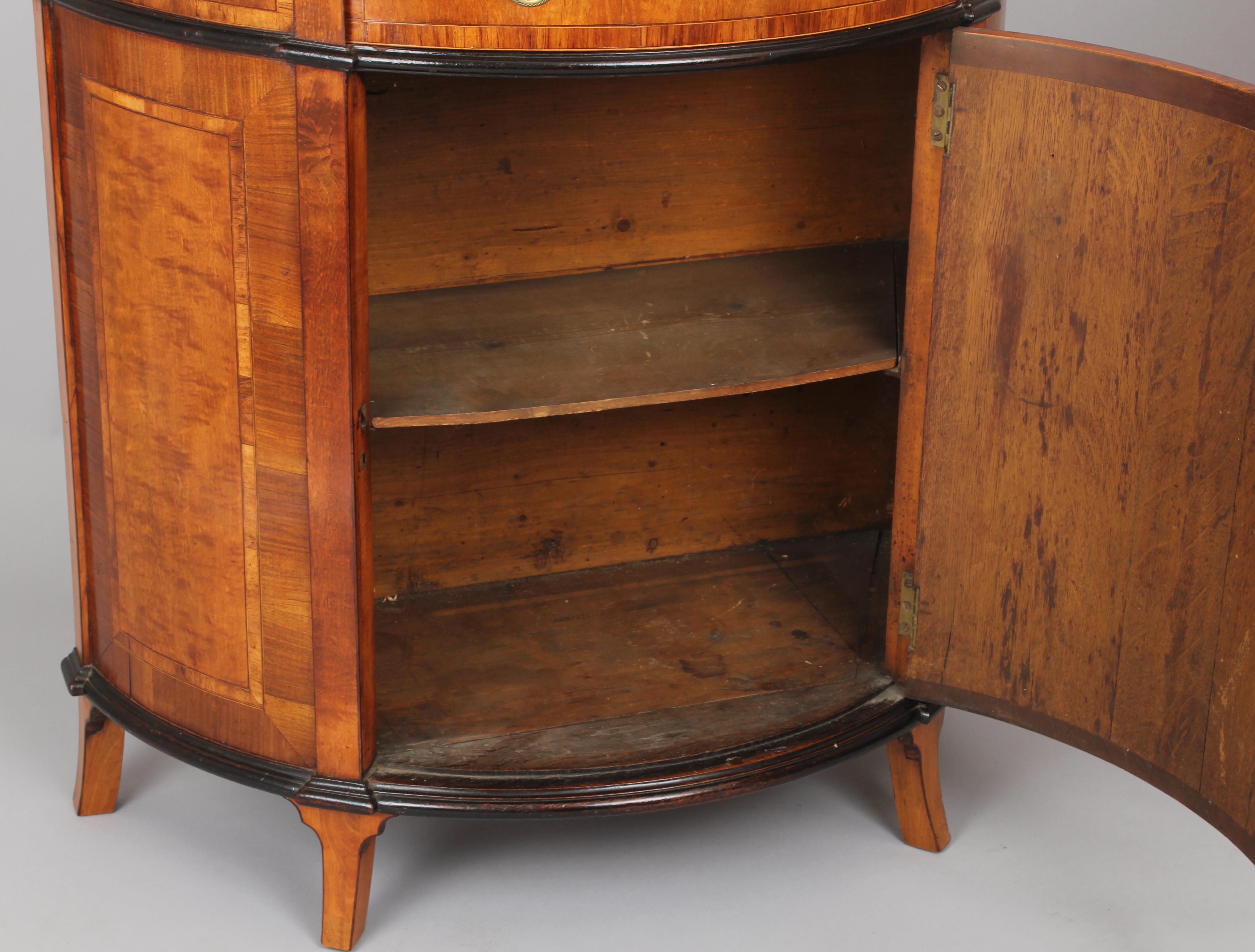 Fine George III period small semi-circular satinwood side-cabinet; the top with a rear overhand and a broad rosewood double-banded border; the shelved cupboard enclosed by a door with an inlaid oval panel, flanked by sides with rectangular panels