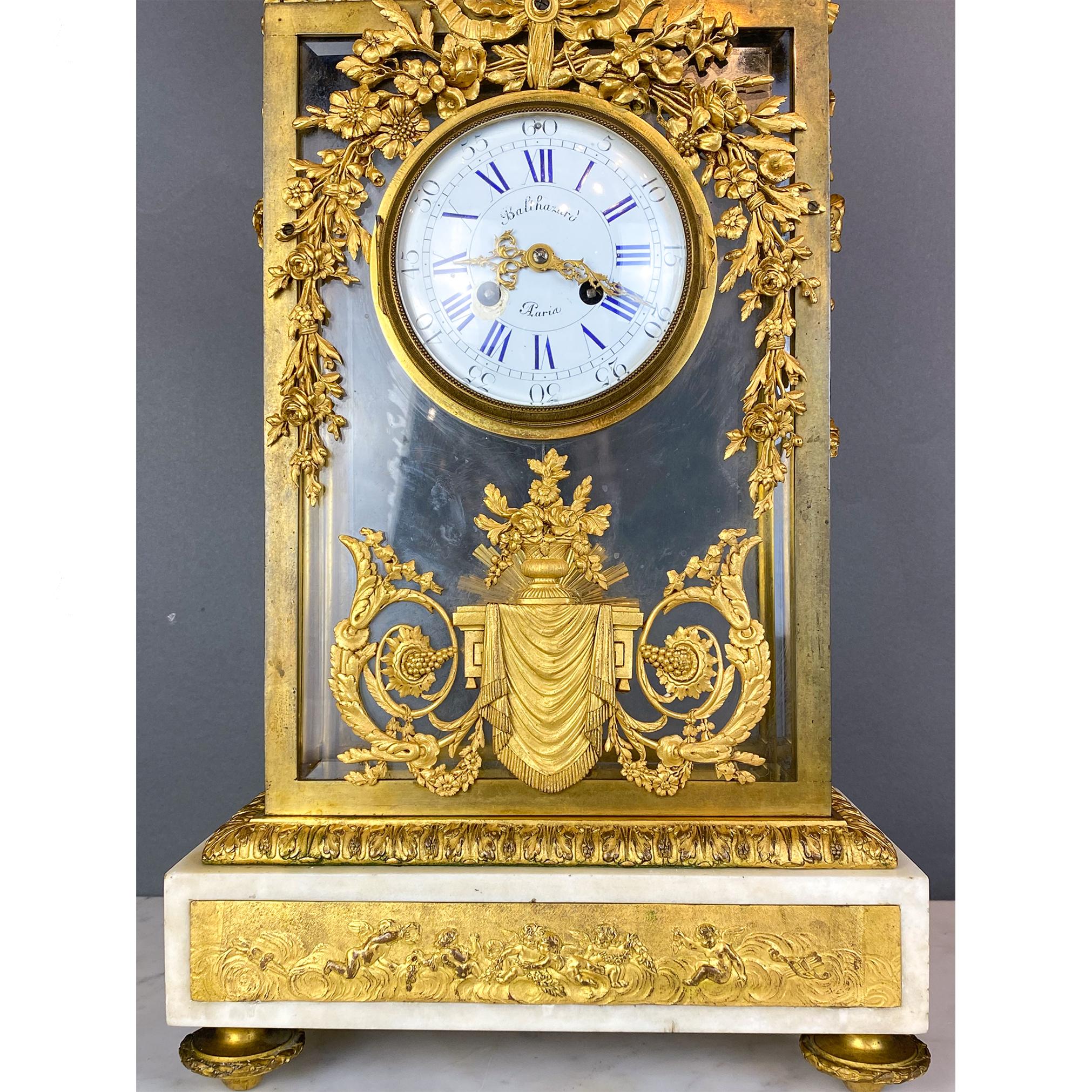 Gilt bronze and glass clock. Rectangular clear glass body showcases an internal sun shaped pendulum. Adorned with foliage, floral and drapery details, the clock is crowned by an urn with an acorn top. The rectangular marble base has a gilt bronze