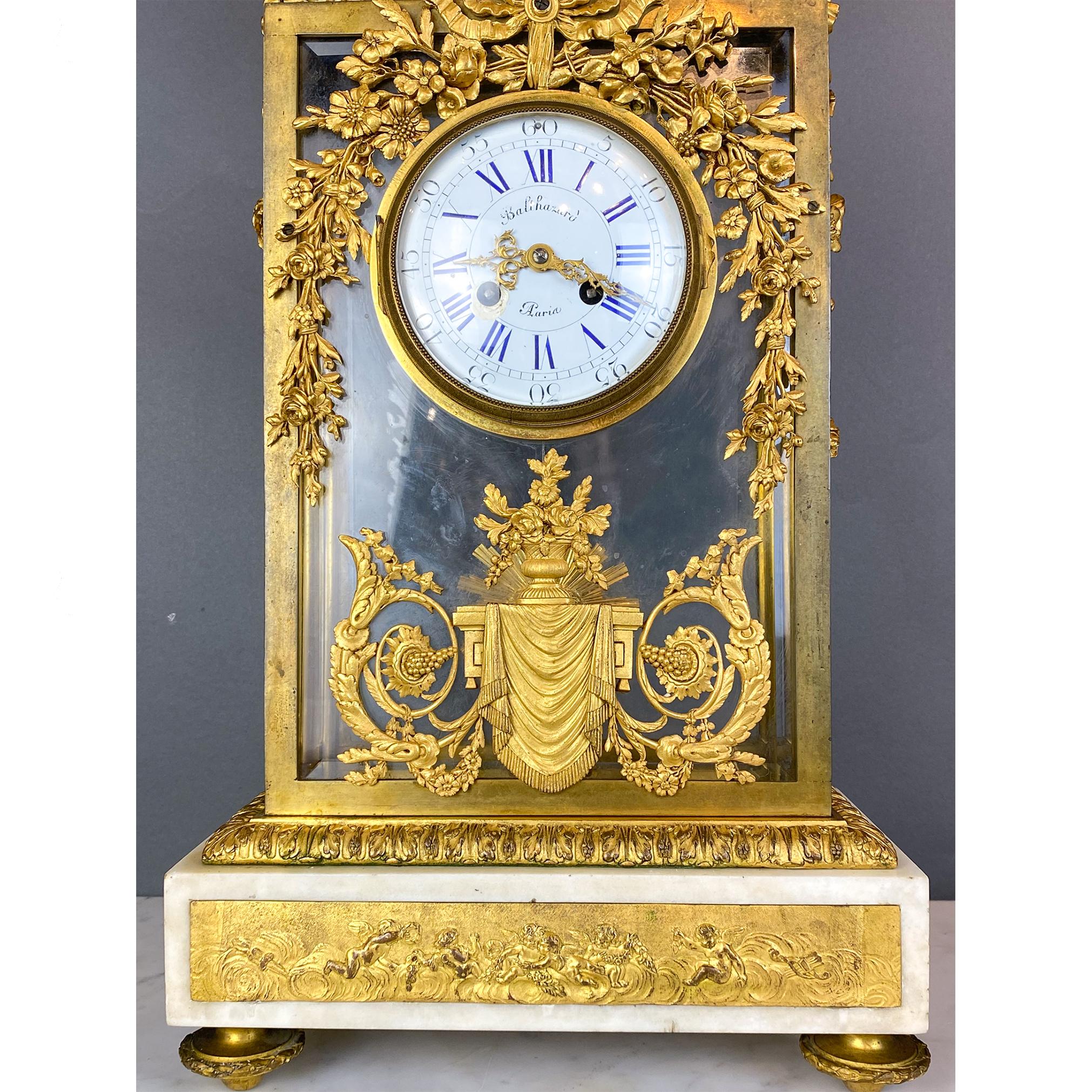 Pendule en bronze doré et verre. Le corps rectangulaire en verre transparent met en valeur un pendule interne en forme de soleil. Ornée de détails de feuillage, de fleurs et de draperies, l'horloge est couronnée d'une urne surmontée d'un gland. La