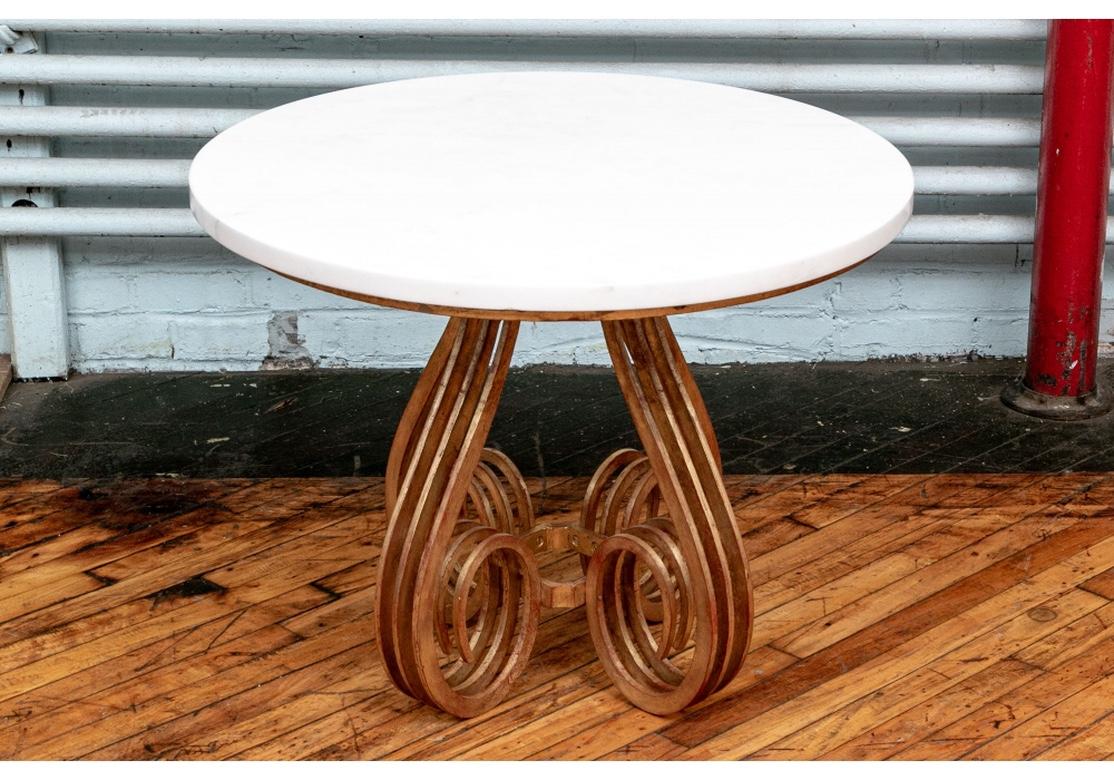 A solidly crafted round iron table with four multiple scrolled supports in gold leaf showing the red sizing beneath. Linked by a square center stretcher on the base. With a round white striated and polished marble top. 
Measures: Diameter 30