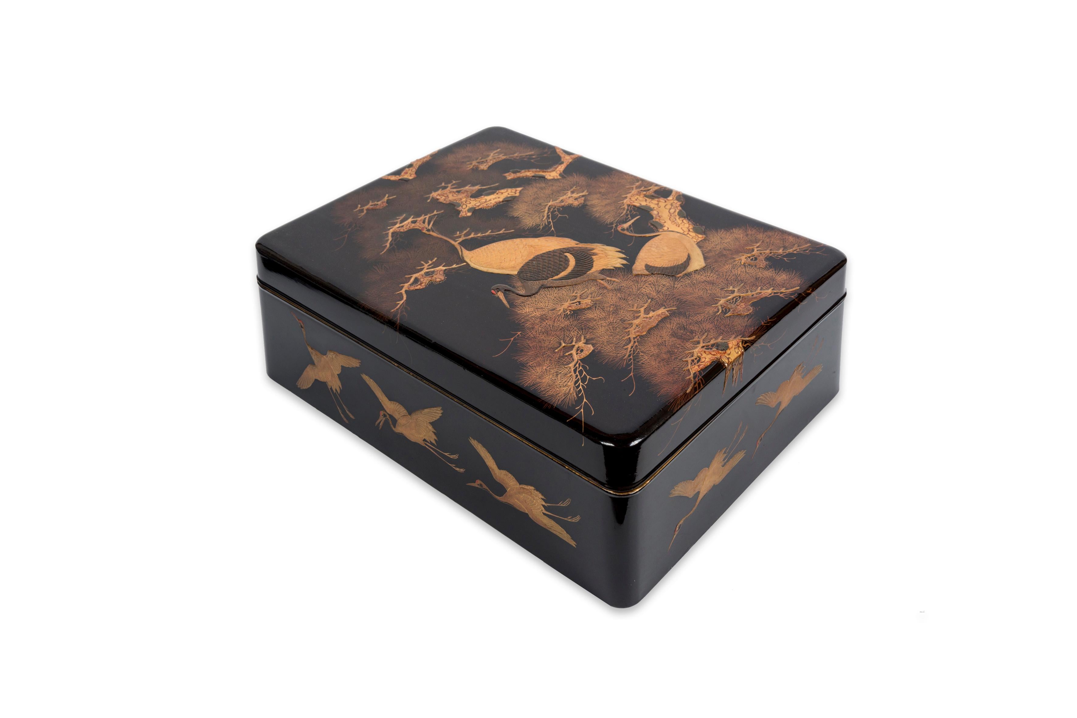 Rectangular paperbox decorated with birds and pine trees.
The lid presents a design mixing several techniques of lacquer work. This is visible with the three cranes represented in the middle of pine branches in gilded maki–e (powdered precious