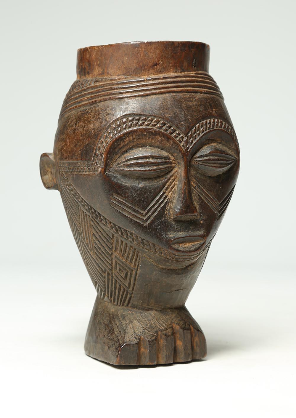 Finely carved wood Kuba cup with expressive face, carefully carved geometric design on the eyebrows and chin, scarification lines across chips. Heavy wear and patina from native use, standing on a carved foot. Early 20th century, Kuba Kingdom,