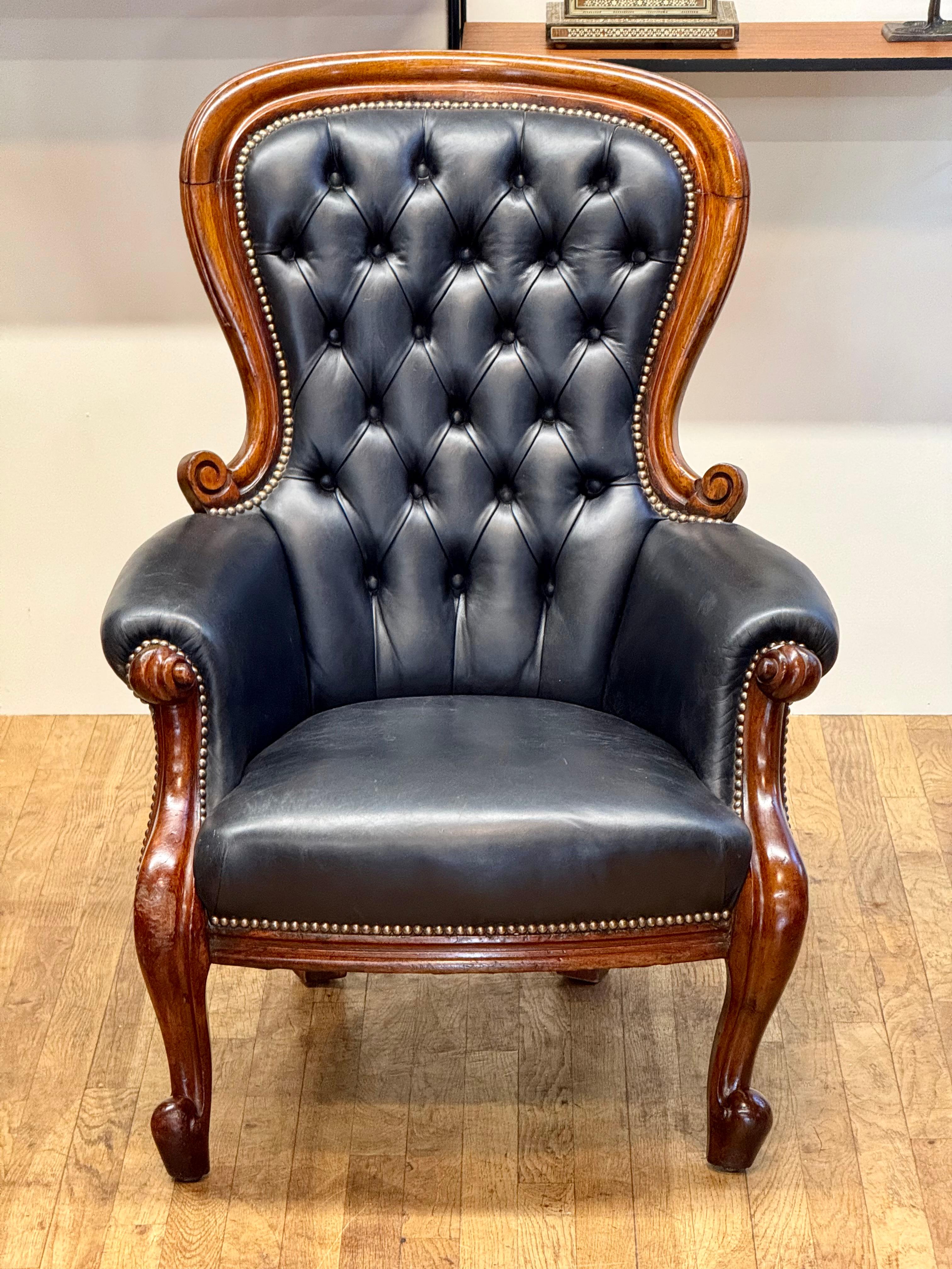 This 19th-century Victorian gentleman's chair is crafted from black leather and mahogany, featuring a tufted back with button detailing for added elegance. The chair's frame is curved and adorned with intricate carvings, showcasing the craftsmanship