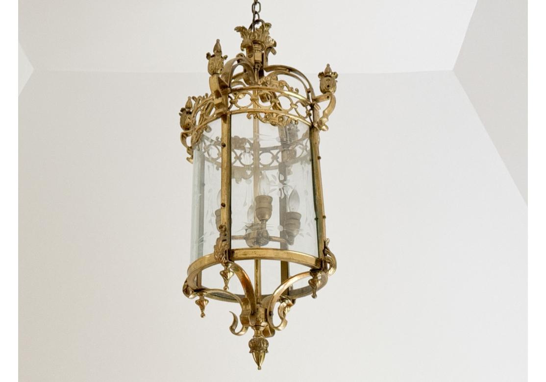 A fine brass and glass chandelier with  French Regency styling, lavishly designed with foliate relief flourishes and torch-like finials on arched supports over an openwork chain motif border heralding a cylindrical glass shade with etched decoration