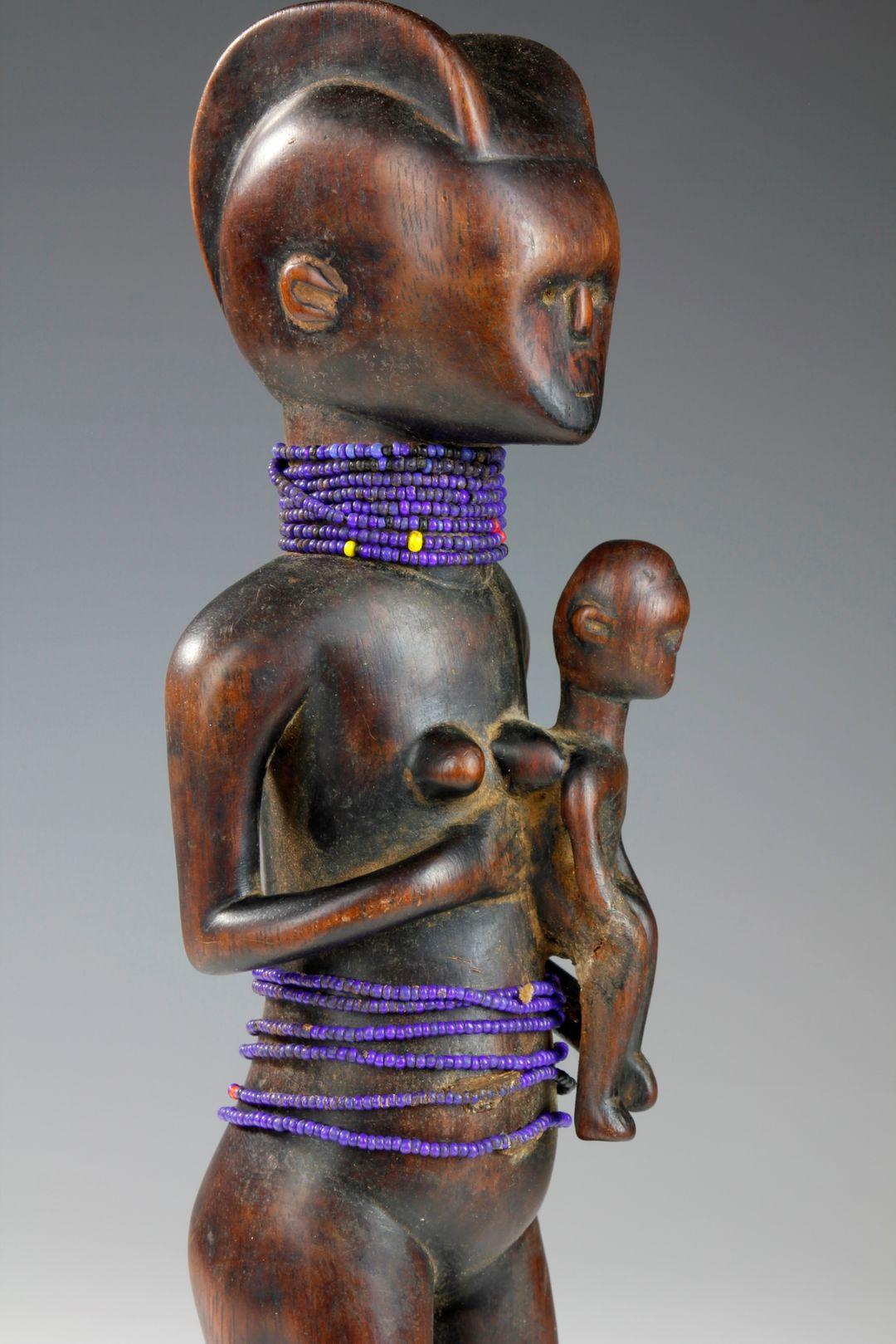 This beautiful mid-twentieth century figurative sculpture depicts a mother, crowned with a sweeping double-parted coiffure, holding her child at her side. Multiple strands of glass blue and red beads adorn the neck and midriff of the mother, and