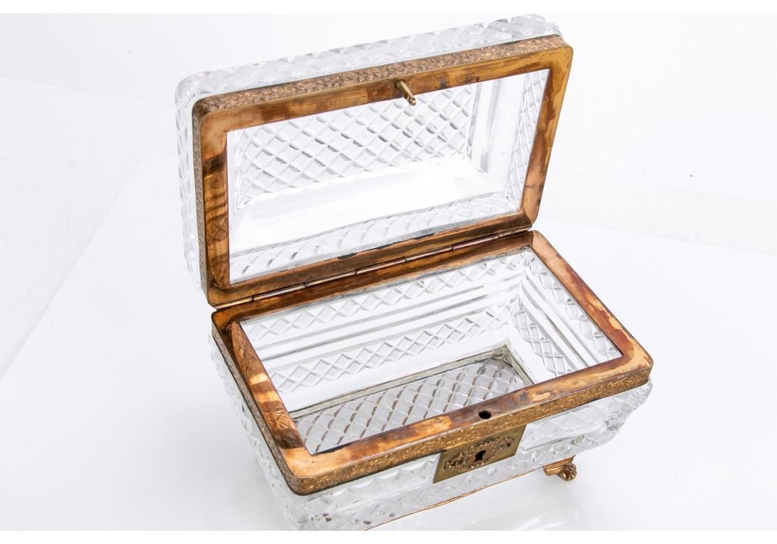 An ormolu mounted crystal box with hinged lid, engraved diamond point cuts, the brass with trailing florals and vines and resting on paw feet. The top tiered with conforming diamond point design. The rectangular escutcheon with relief