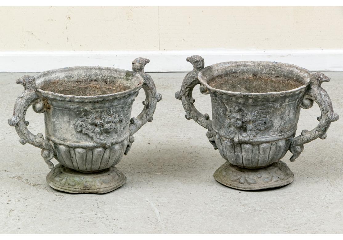Antique pair of lead garden urns each featuring an urn raised on a rounded pedestal, raised cherub motif and c-scroll handles wrapped with mythological creatures. Each with drainage holes to bottom.

Dimensions:  20