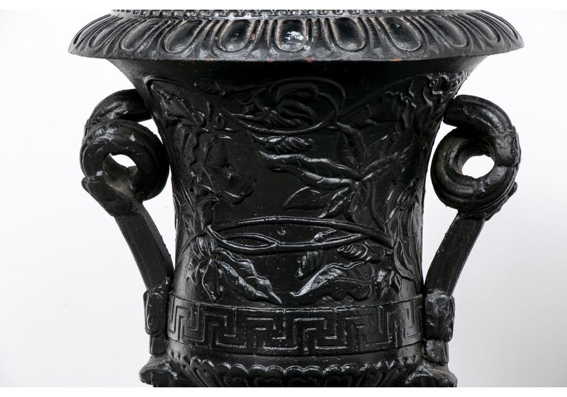 The iron urn pair having superlative overall flowering vine body decoration. Each urn having egg and dart motif rims over the vine designs. The bases are Greek key banded and having scrolled handles terminating on lower satyr masks. The pair are