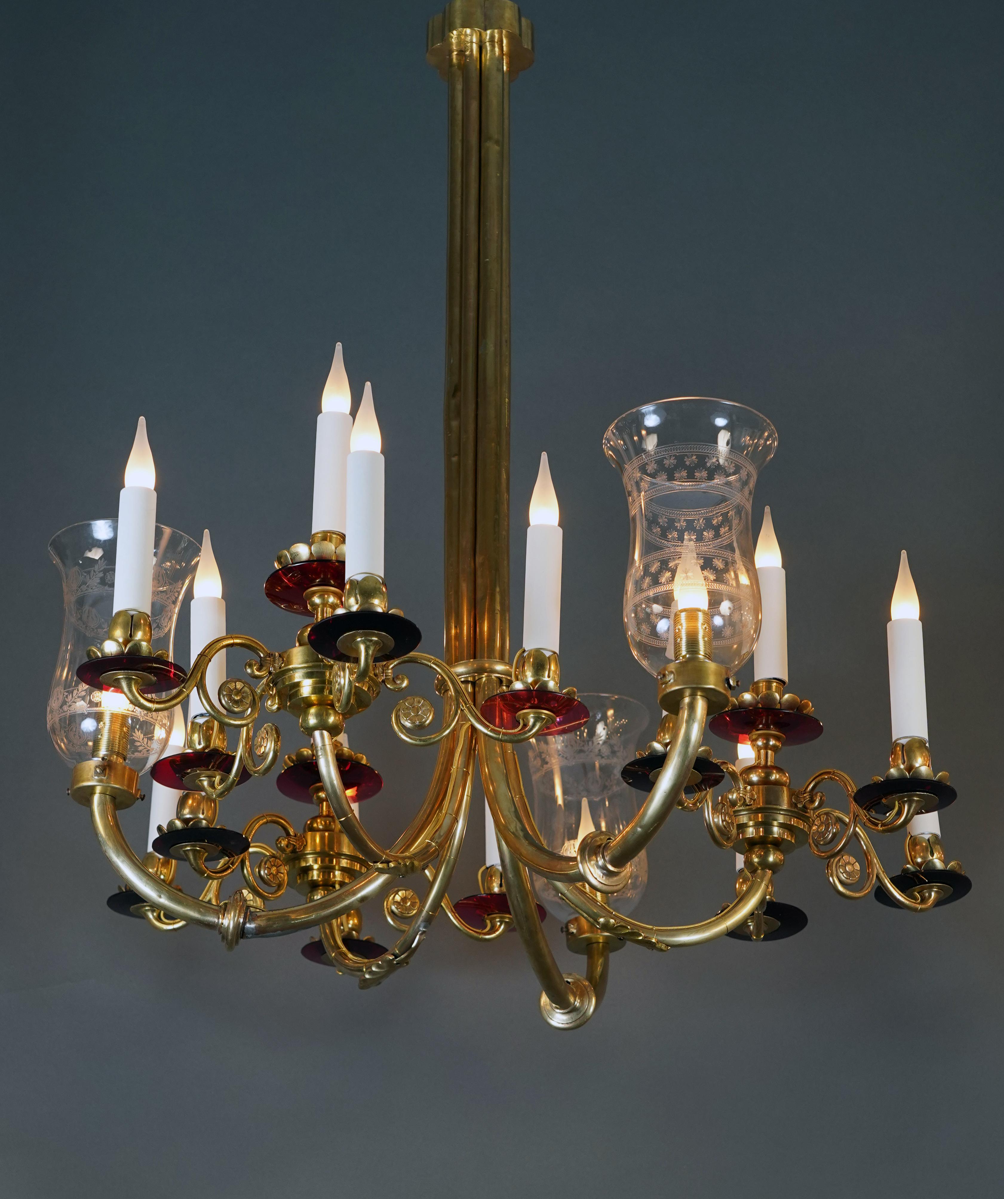 Elegant pair of brass and glass chandeliers whose shafts are formed by the extensions of six light arms joined together in ringed columns.
The light arms, with their curved lines underlined by stylized flowers and palms, end in red glass circles