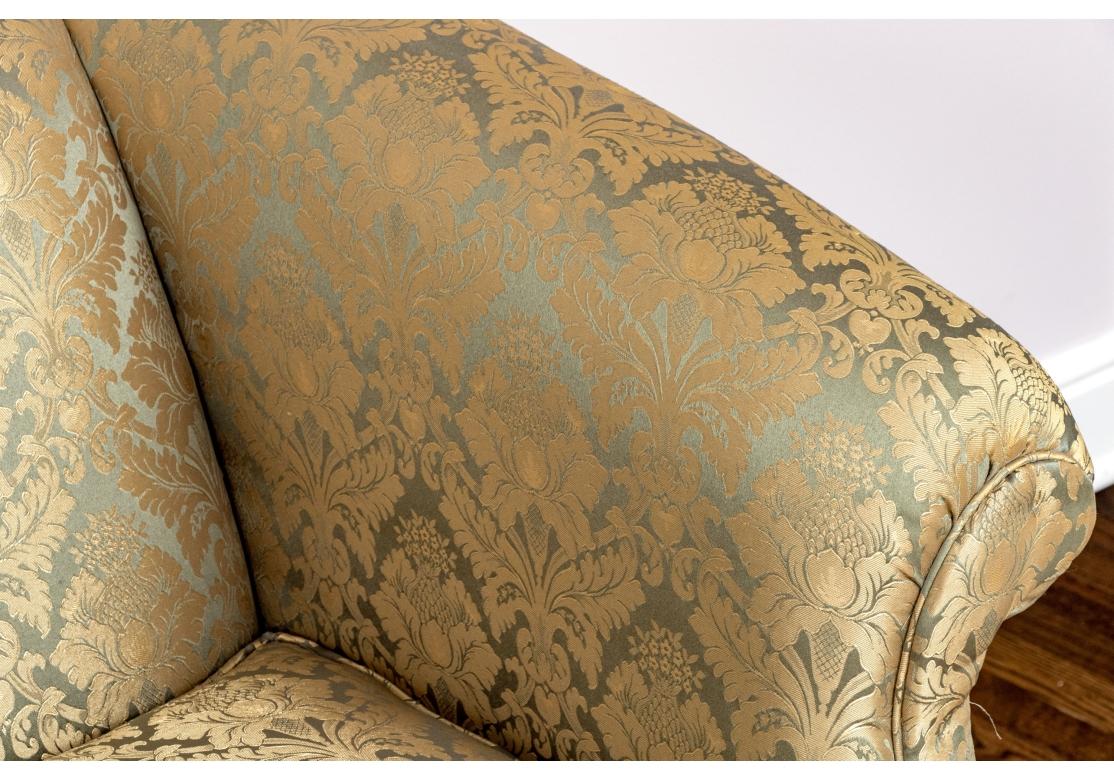 Lavishly upholstered with gold floral motifs over a shimmery blue ground. The sofa with skirt to conceal the underneath, two loose upholstered seat cushions with piped trim. Rolled arms and Classic Camelback form.   

Dimensions: 62 1/4