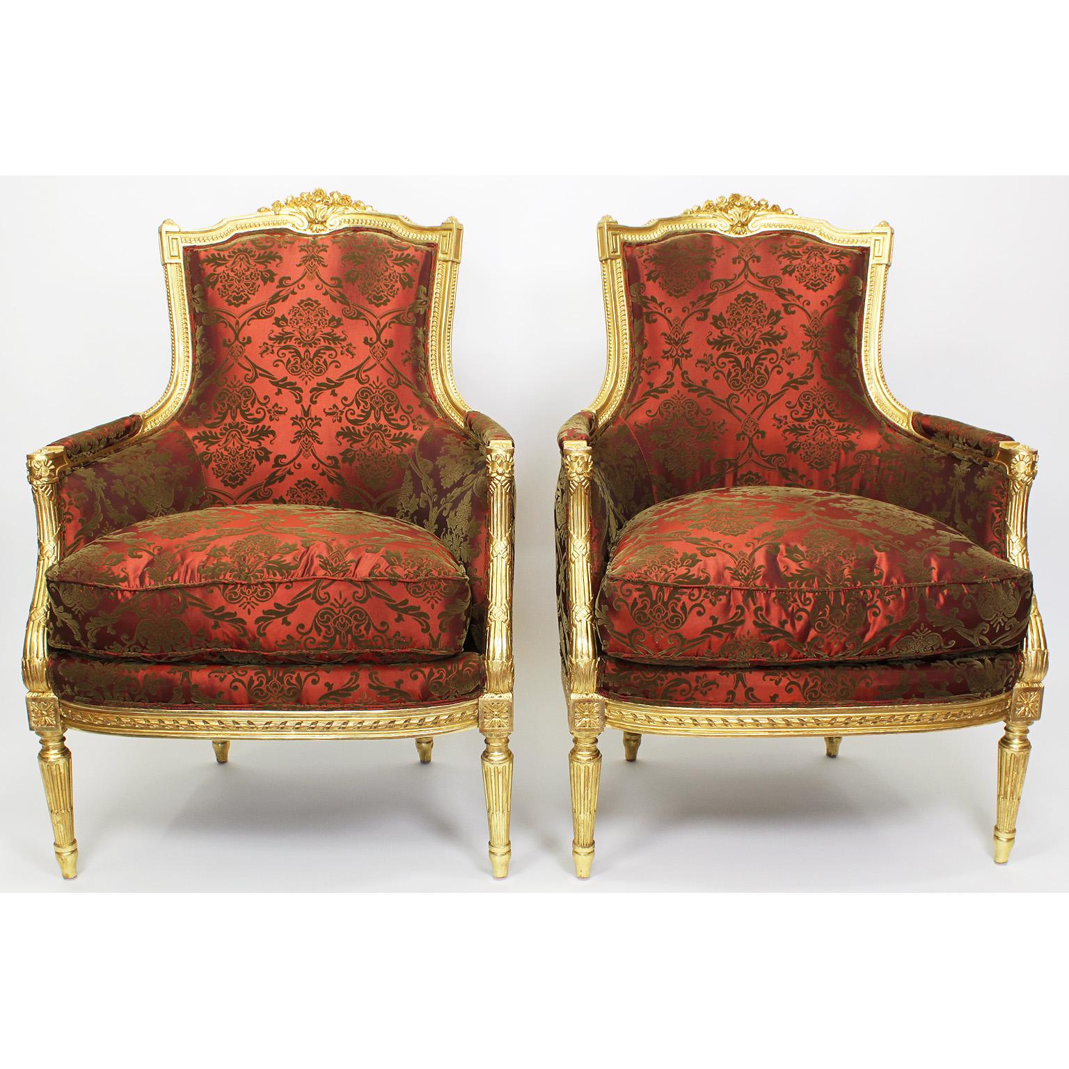 A fine pair of French 19th-20th century Louis XVI style giltwood carved bergères, the recently upholstered and restored armchairs with a padded back, sides and loose cushion seat in floral damask, crowned with a floral carving and raised on four