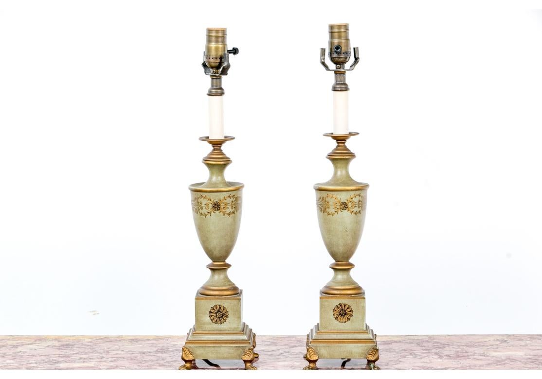 Coppia di lampade a urna dipinte in Celadon polveroso con decorazioni a foglie d'acanto stilizzate e accenti dorati. Le basi presentano decorazioni floreali e hanno basi quadrate che poggiano su piedi a zampa. Su ogni base c'è un'iscrizione