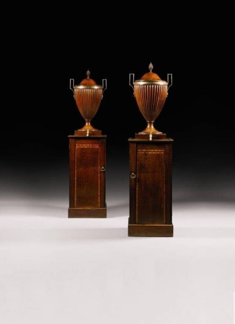 Each cistern is in the form of a fluted vase on a rectangular pedestal. The lead-lined vases have brass handles, removable lids with pineapple finials and taps. The crossbanded pedestals have cupboard doors enclosing slatted shelves for warming