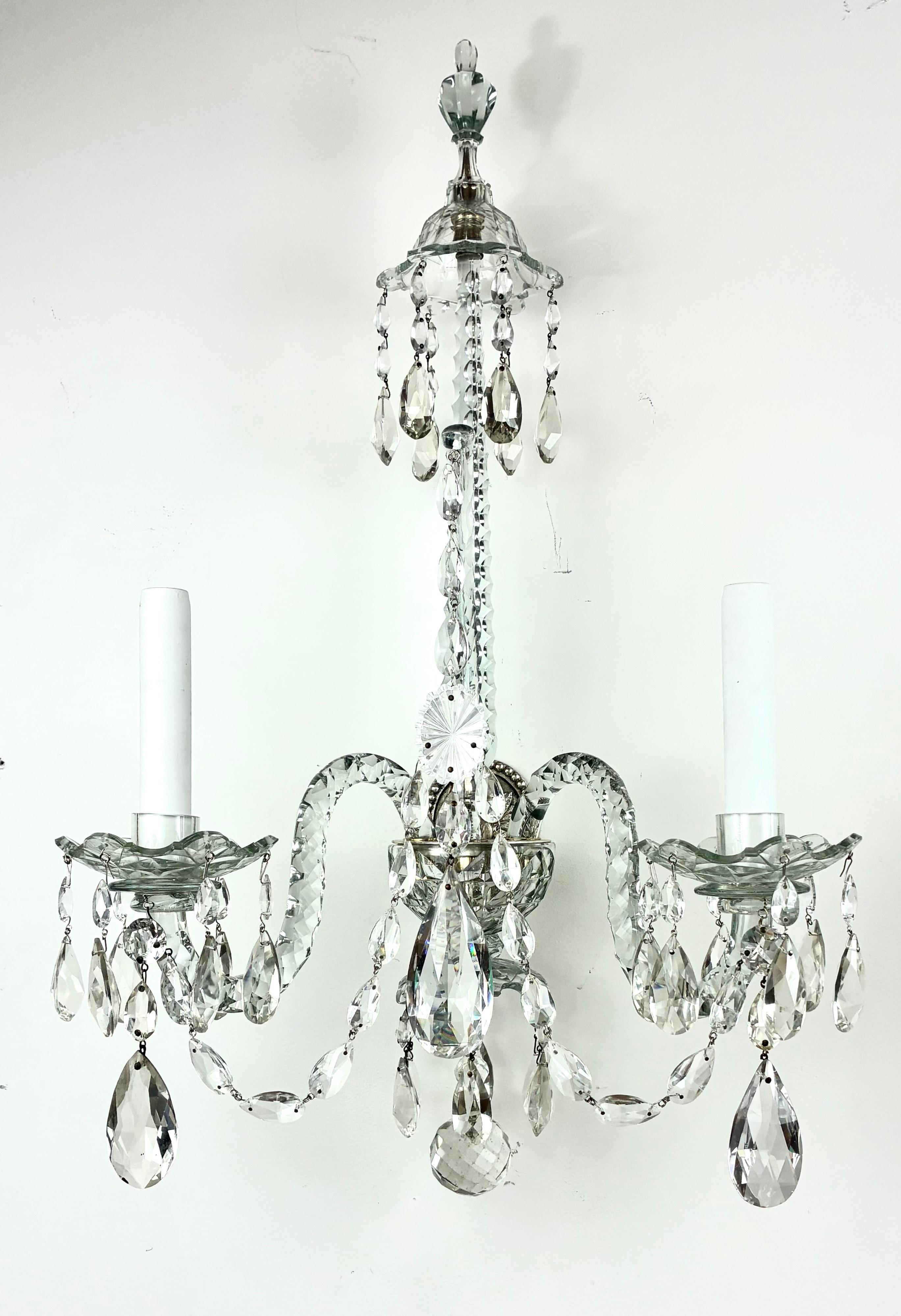 This fine and impressive pair of George III style two light sconces are truly exceptional.
The candle arms are fitted with Van Dyke style crystal nozzles and bobeches. The almond shaped swag chain is suspended from S form “snake scrolls” terminating