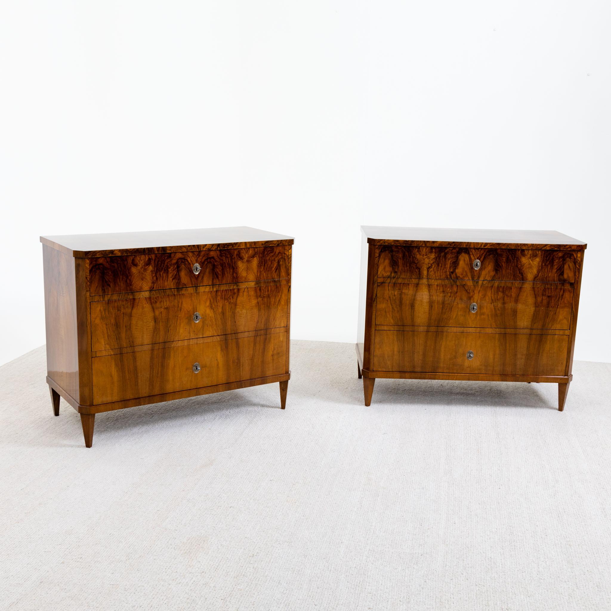 Pair of Italian Biedermeier chests of drawers featuring three drawers each, standing on square pointed legs. The bodies, with beveled corners, are veneered in walnut and meticulously hand-polished. The drawers are adorned with brass escutcheons,