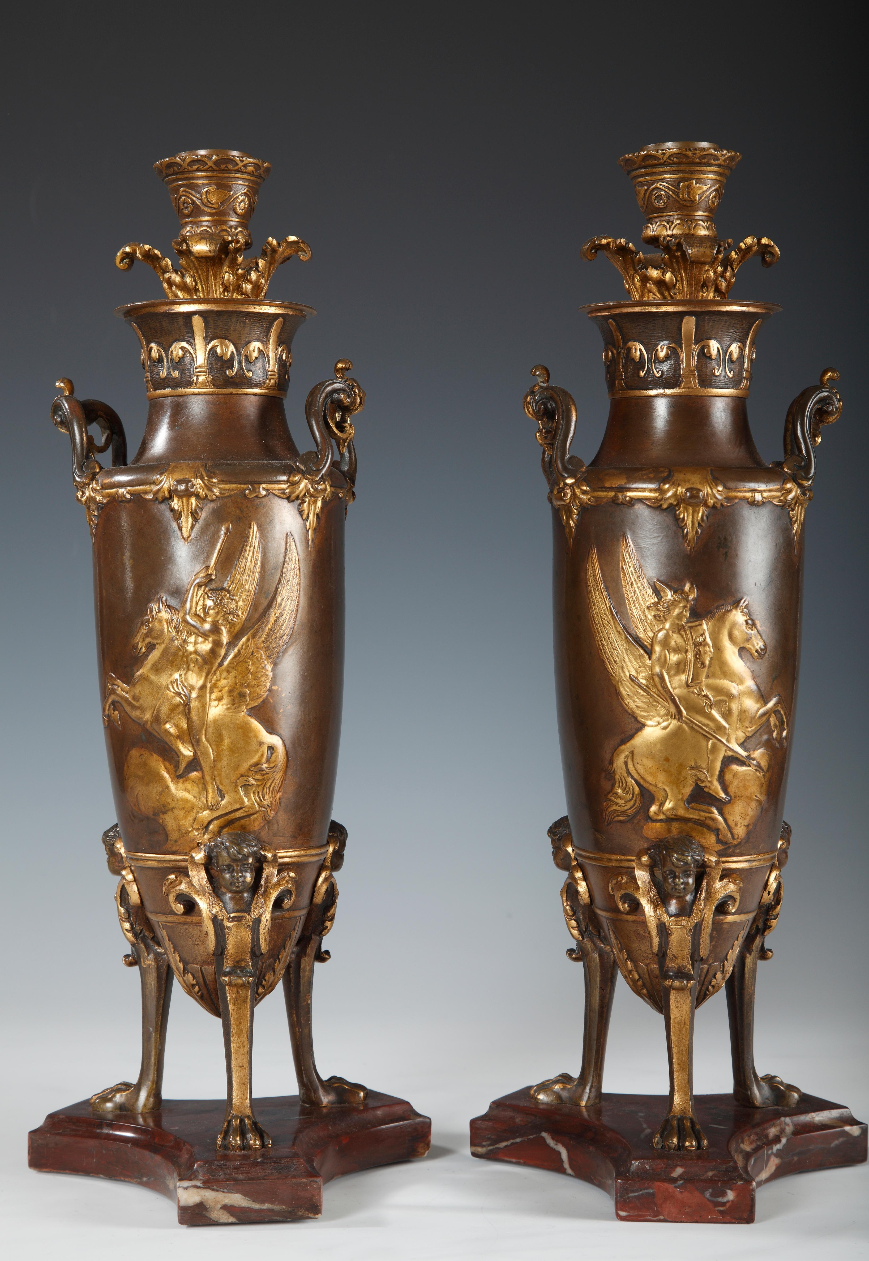 Paire de chandeliers de style grec en forme d'amphore en bronze doré et patiné attribuée à F. Levillain et F. Barbedienne. Chacune est flanquée de deux poignées et décorée de cavaliers en relief. Elle repose sur trois pieds de lion coiffés de bustes